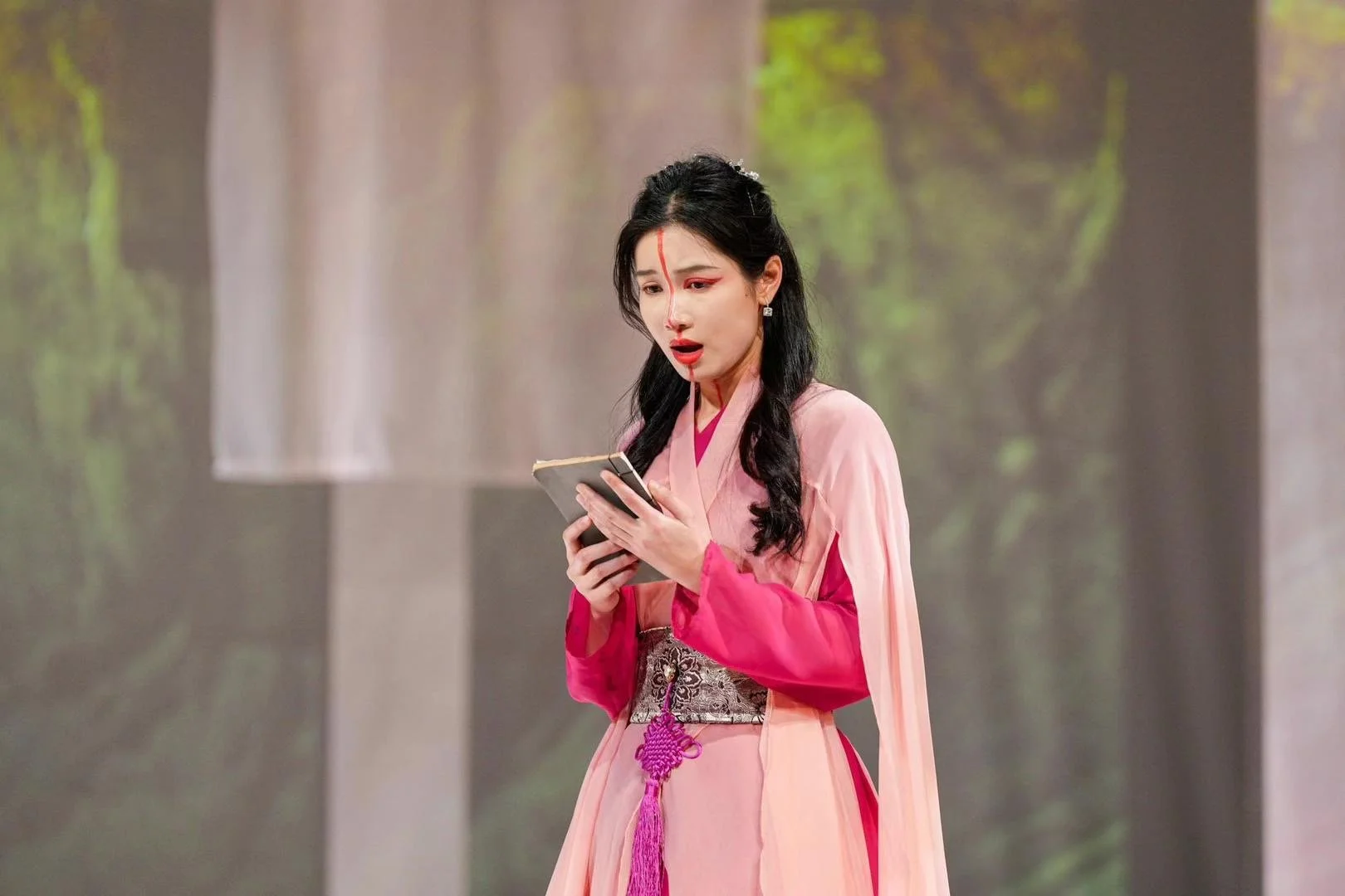 A woman dressed in traditional pink and red clothing, with long black hair, holds a small book or notebook and appears surprised or shocked, standing against a blurred background with trees.