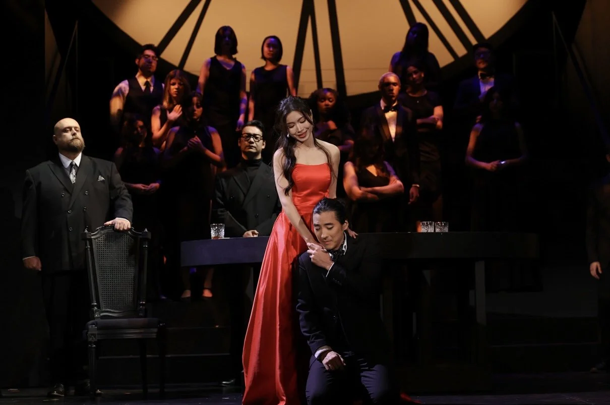 A stage scene with a woman in a red dress and black-haired man in a black suit, surrounded by a choir in formal black attire, with some on a platform and others standing behind. The woman is holding the man's shoulders as he kneels, in an emotional m
