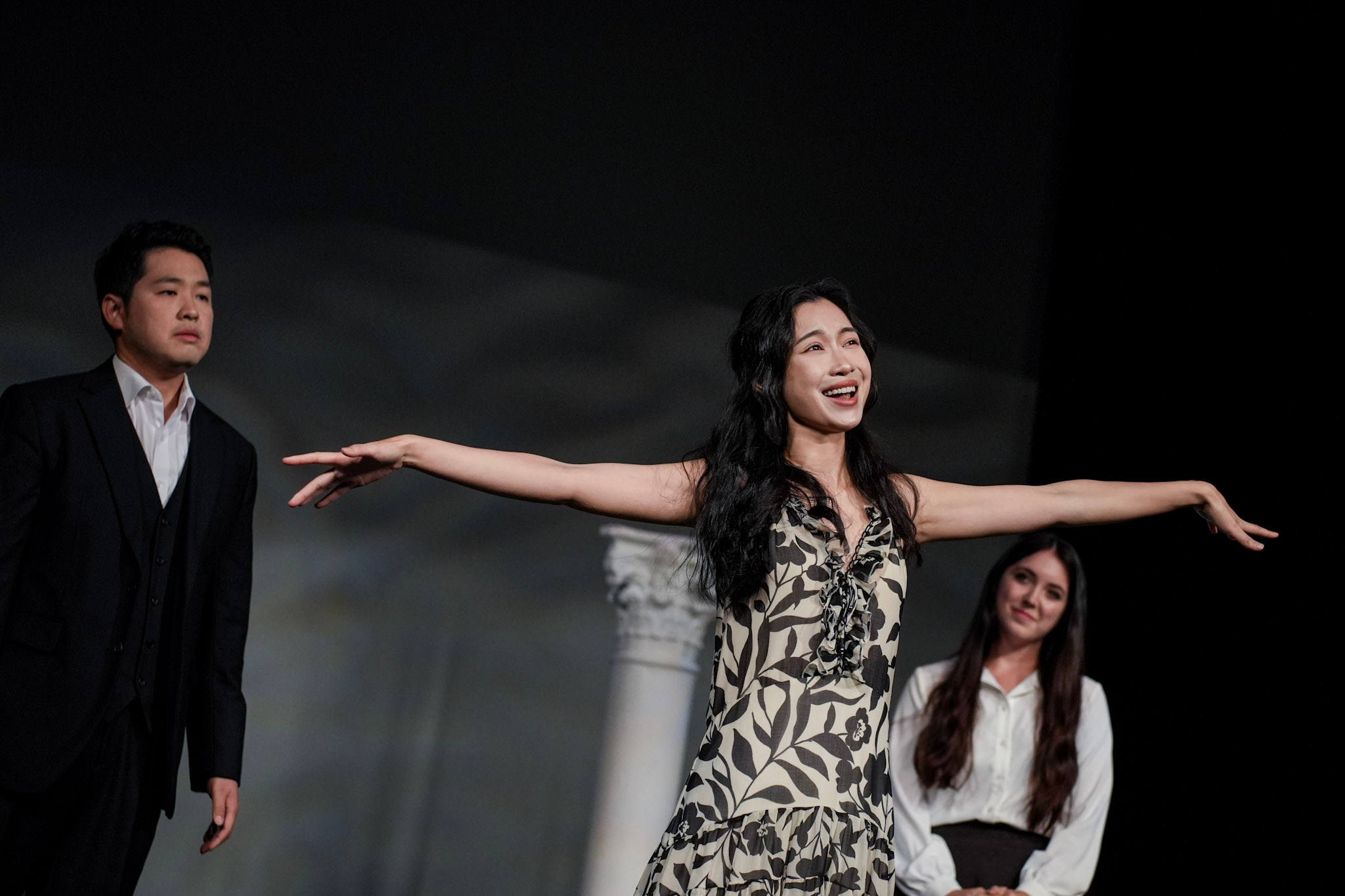 Group of women and a man on stage. One woman is smiling and extending her arms, wearing a patterned dress. The man is in a black suit with a serious expression, standing to her left. Other women are dressed in white and appear to be listening.