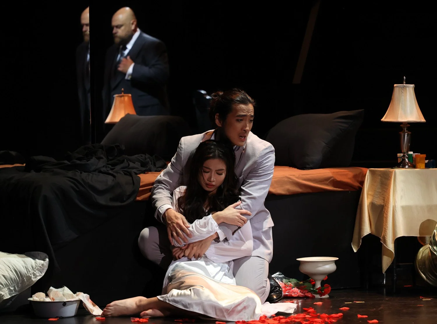 A woman sitting on the floor in front of a bed, hugging another woman who appears distressed. Two men in suits stand in the background with serious expressions. The scene appears intense and emotional, with a dramatic setting including scattered obje