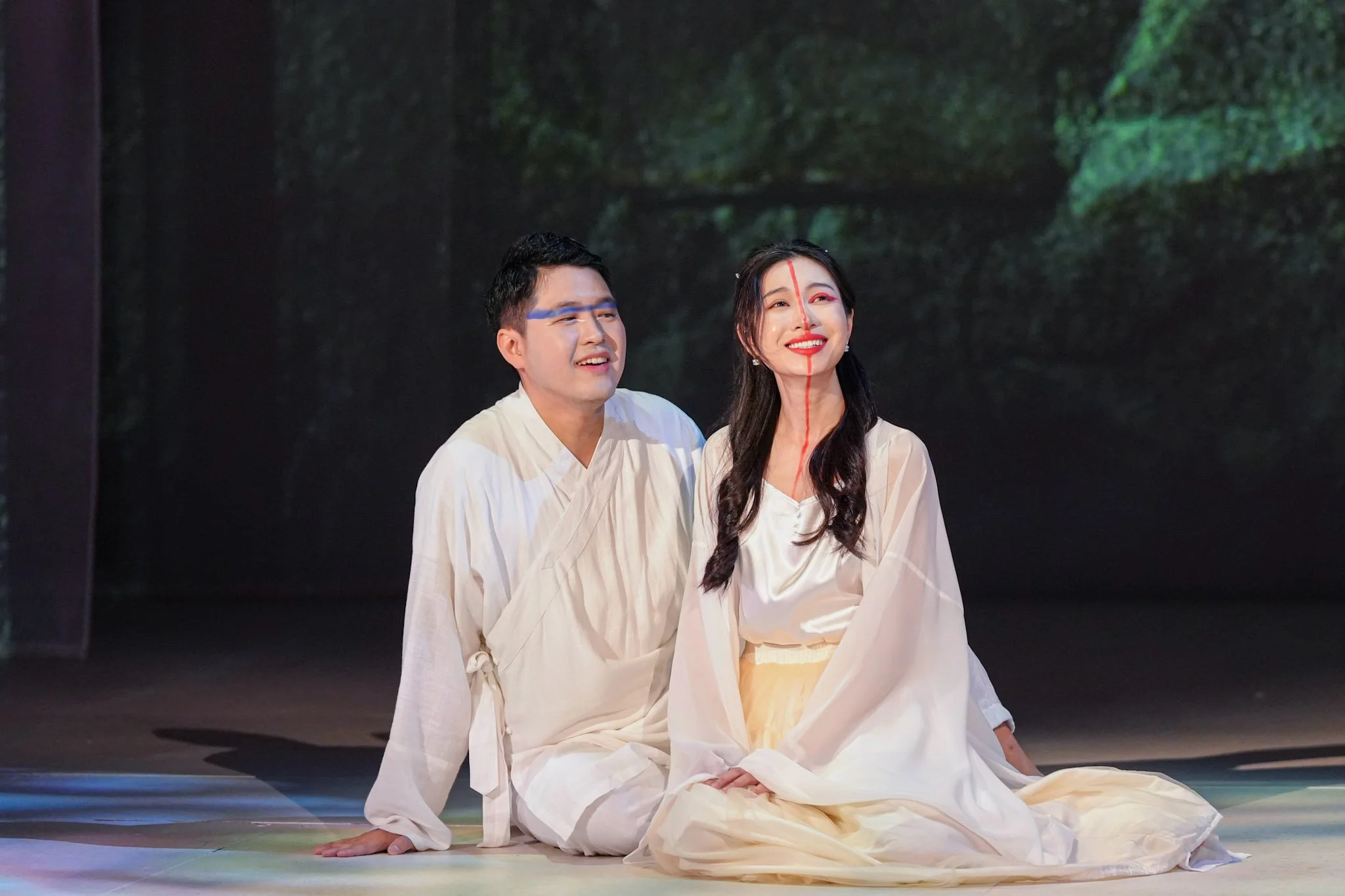 A man and a woman dressed in white, sitting on stage with a dark background, possibly performing in a theatrical production.