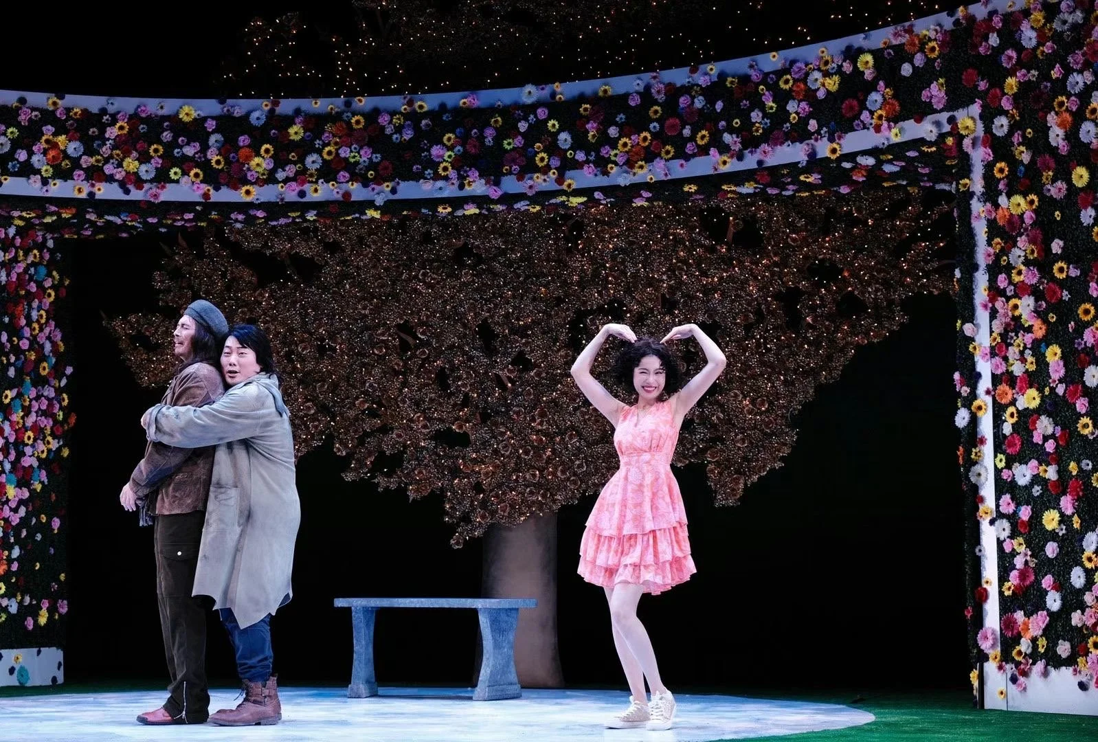 Three people on a stage with a floral backdrop. Two people are standing close together on the left, hugging, while a woman in a pink dress stands to the right, smiling and making a heart shape with her arms above her head.