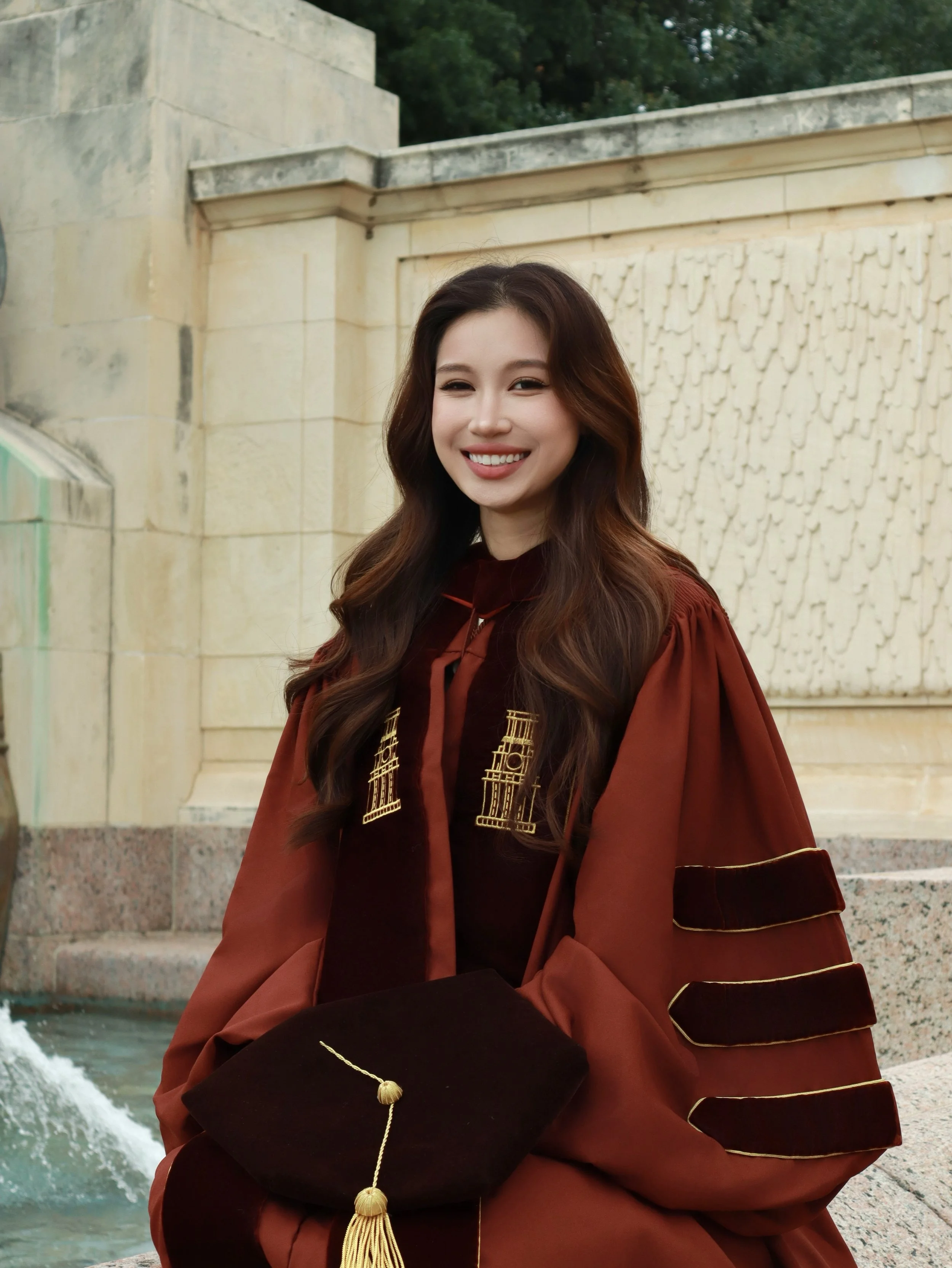 A young woman with long brown hair smiling, dressed in a maroon graduation gown and holding a black cap with gold embroidery, sitting outdoors with a stone wall and fountain in the background.