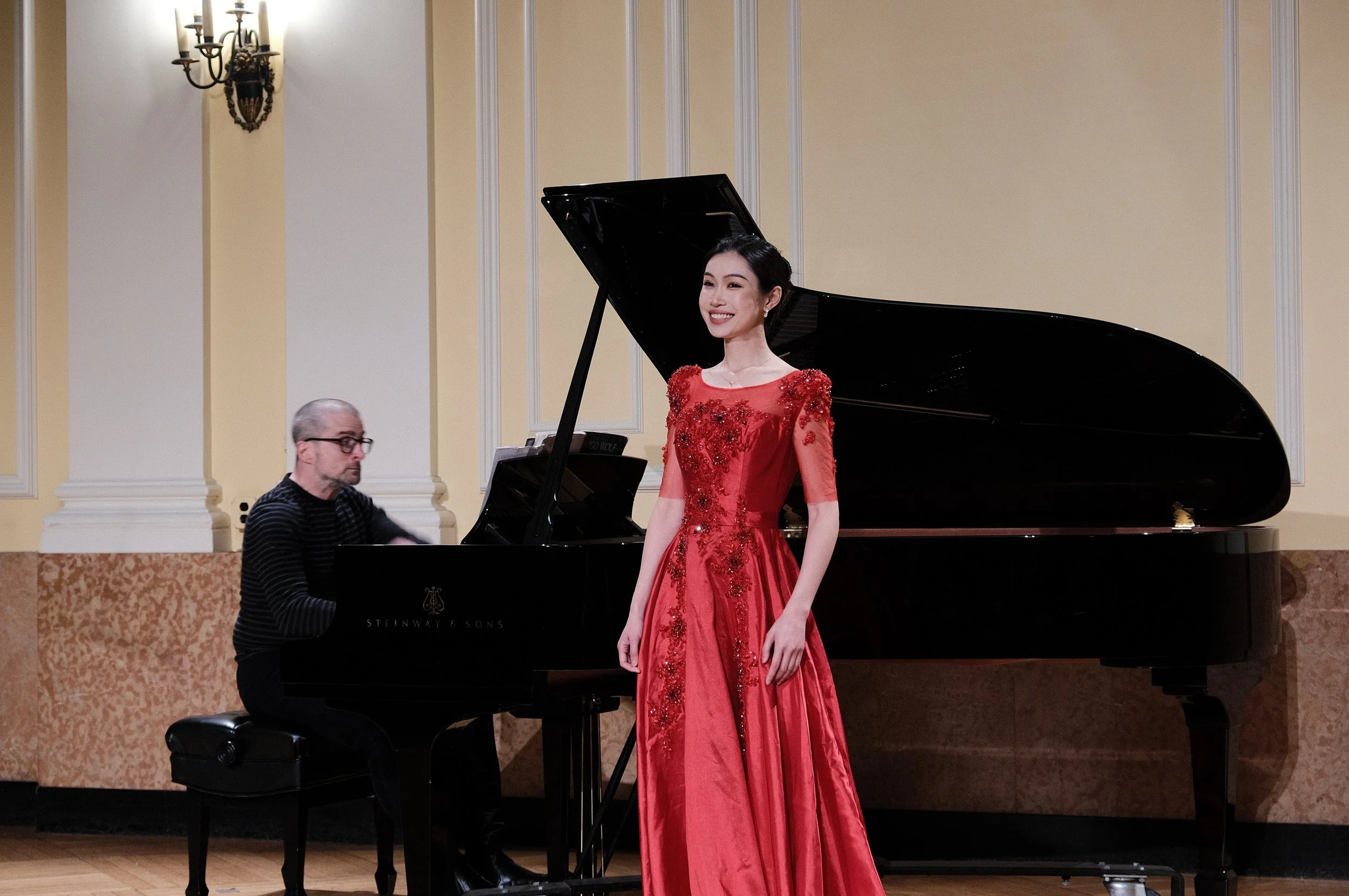 A woman in a red dress standing next to a man playing a grand piano, in a formal indoor setting with beige walls and a chandelier.