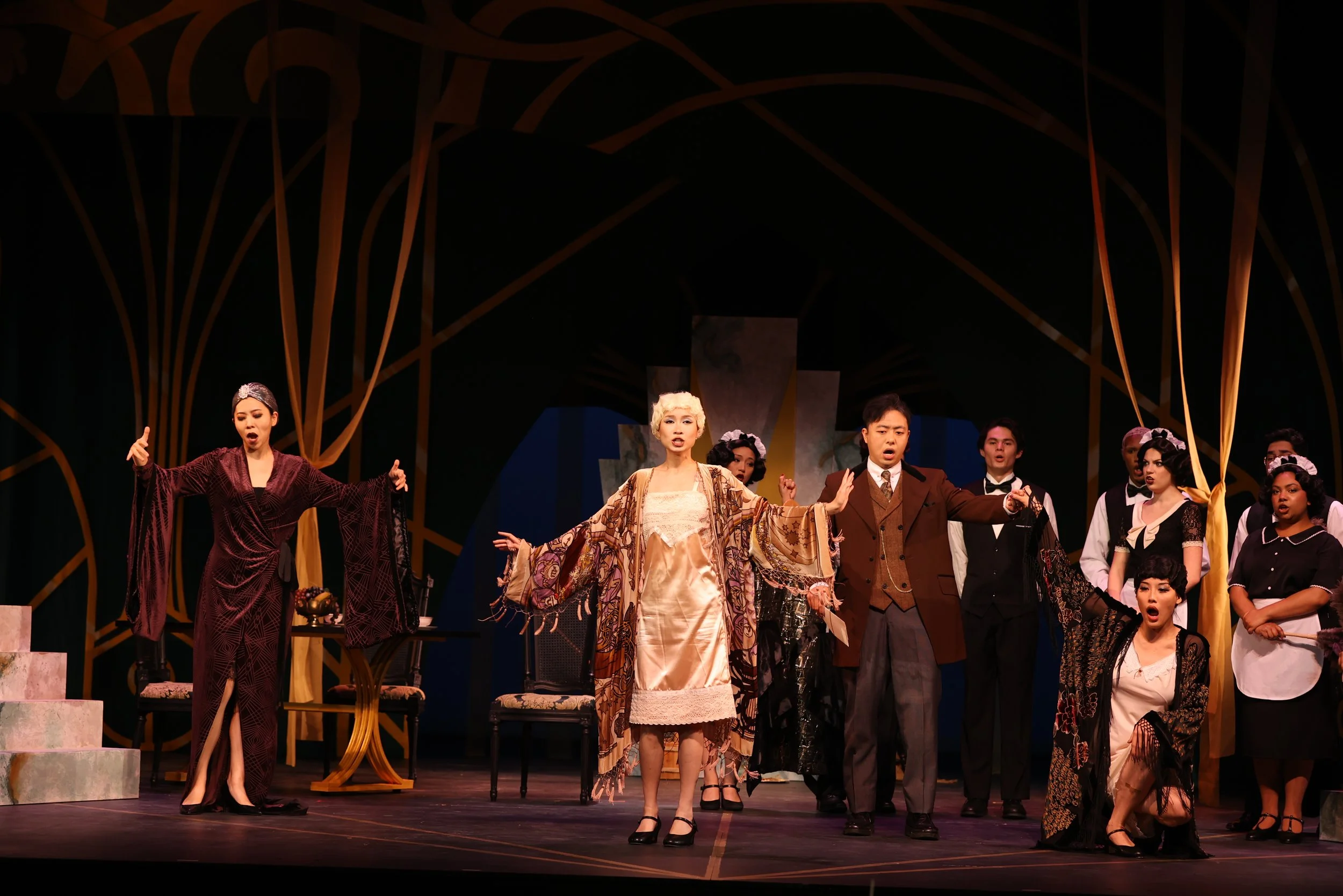 The image shows a cast of theatrical performers on stage, dressed in period costumes, standing with their arms outstretched, likely at the end of a performance or scene, with a decorative stage backdrop behind them.