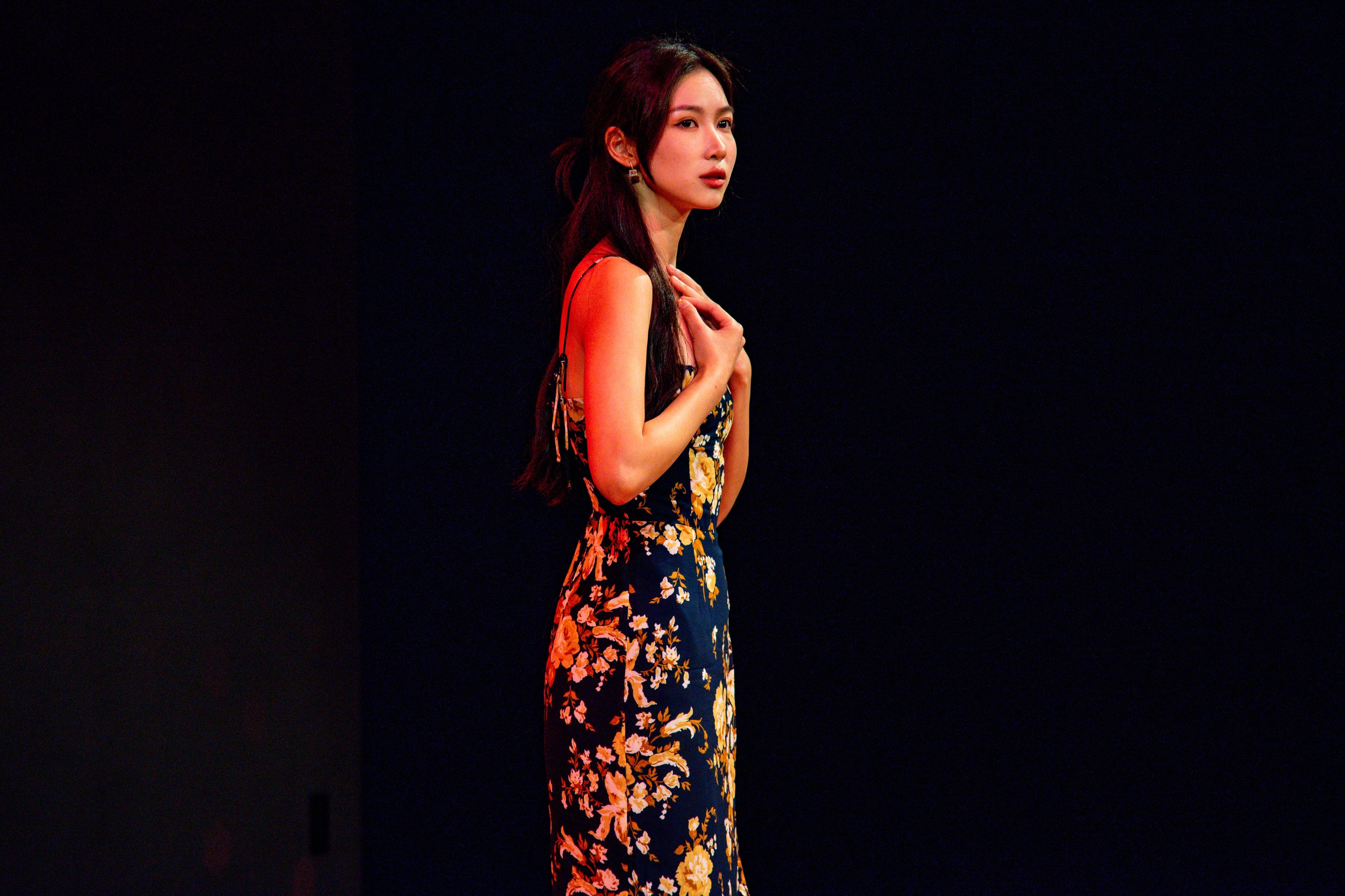 A woman with long dark hair in a floral dress with orange and yellow flowers, standing against a black background.