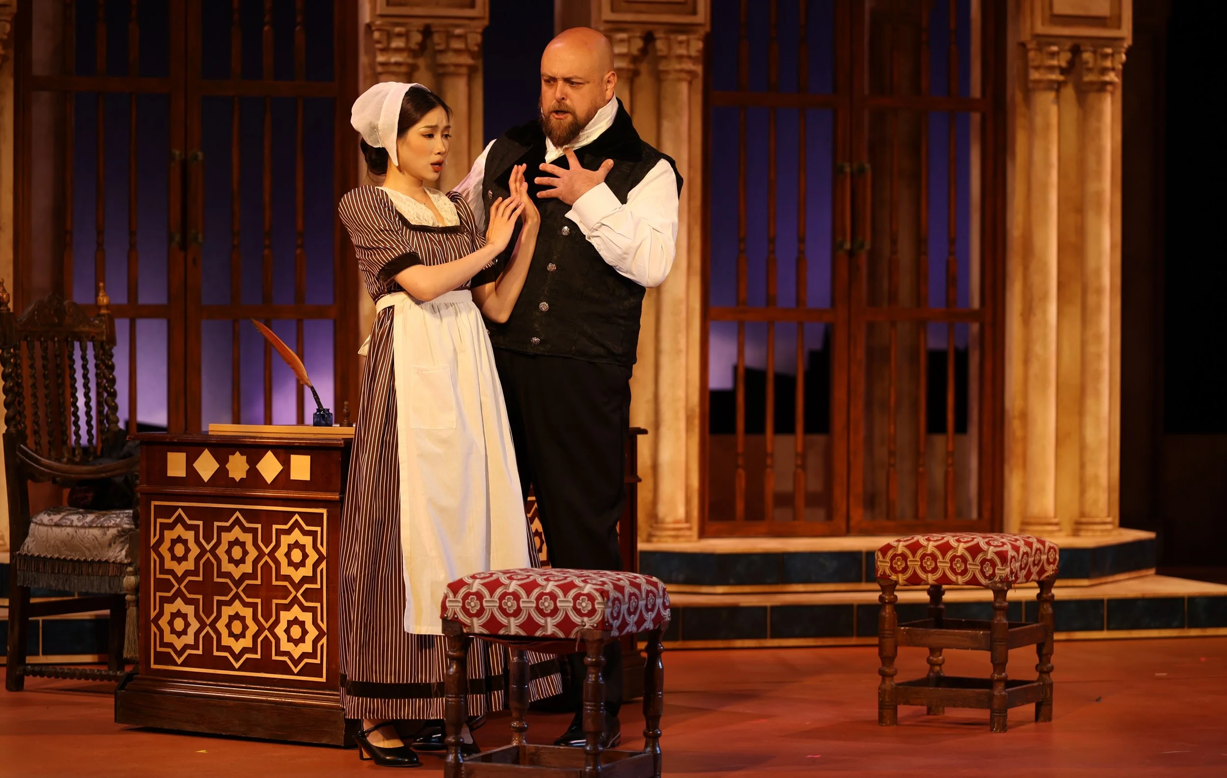 Two actors in historical costumes perform on stage with a wooden set, including chairs and decorative panels, creating the scene of a theatrical play.