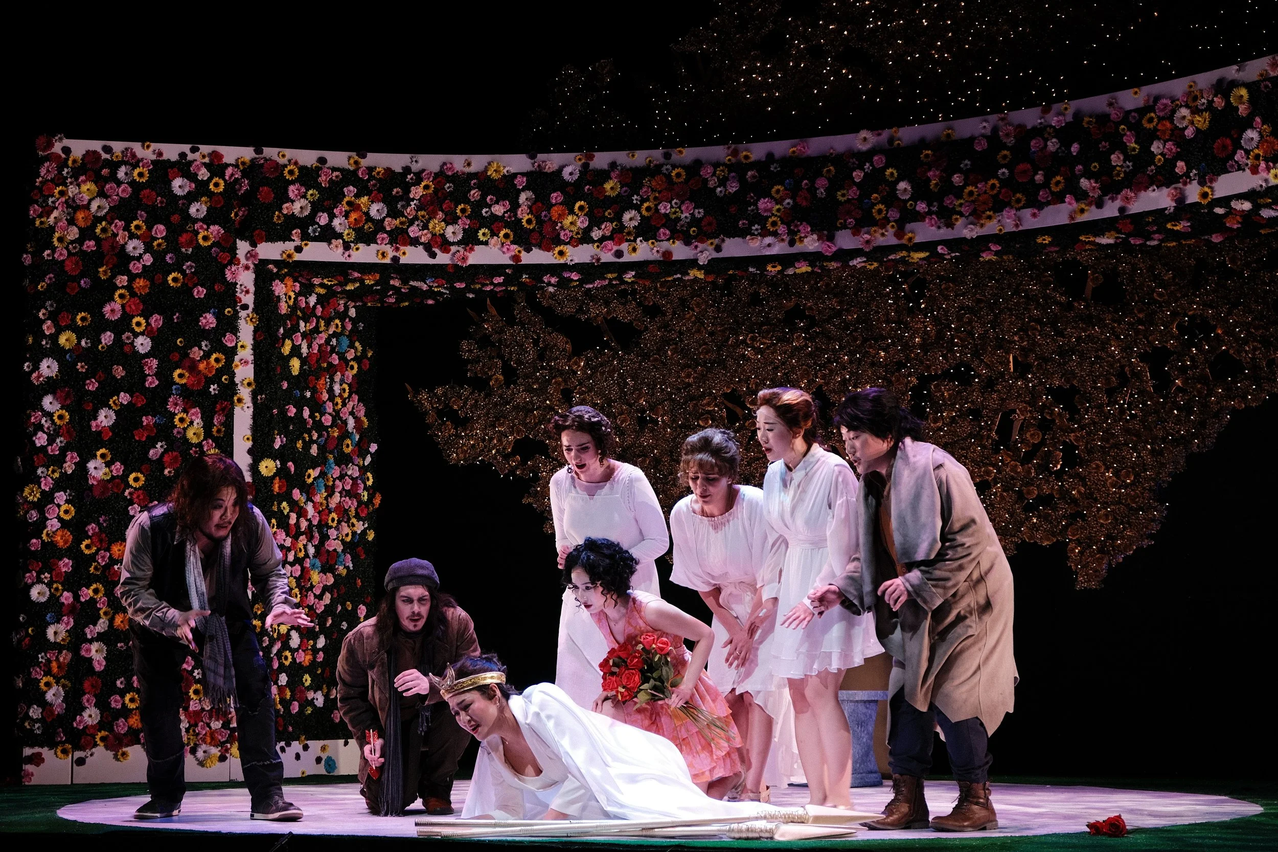 A group of actors on stage performing a scene, with a backdrop decorated with flowers and illuminated by stage lighting.
