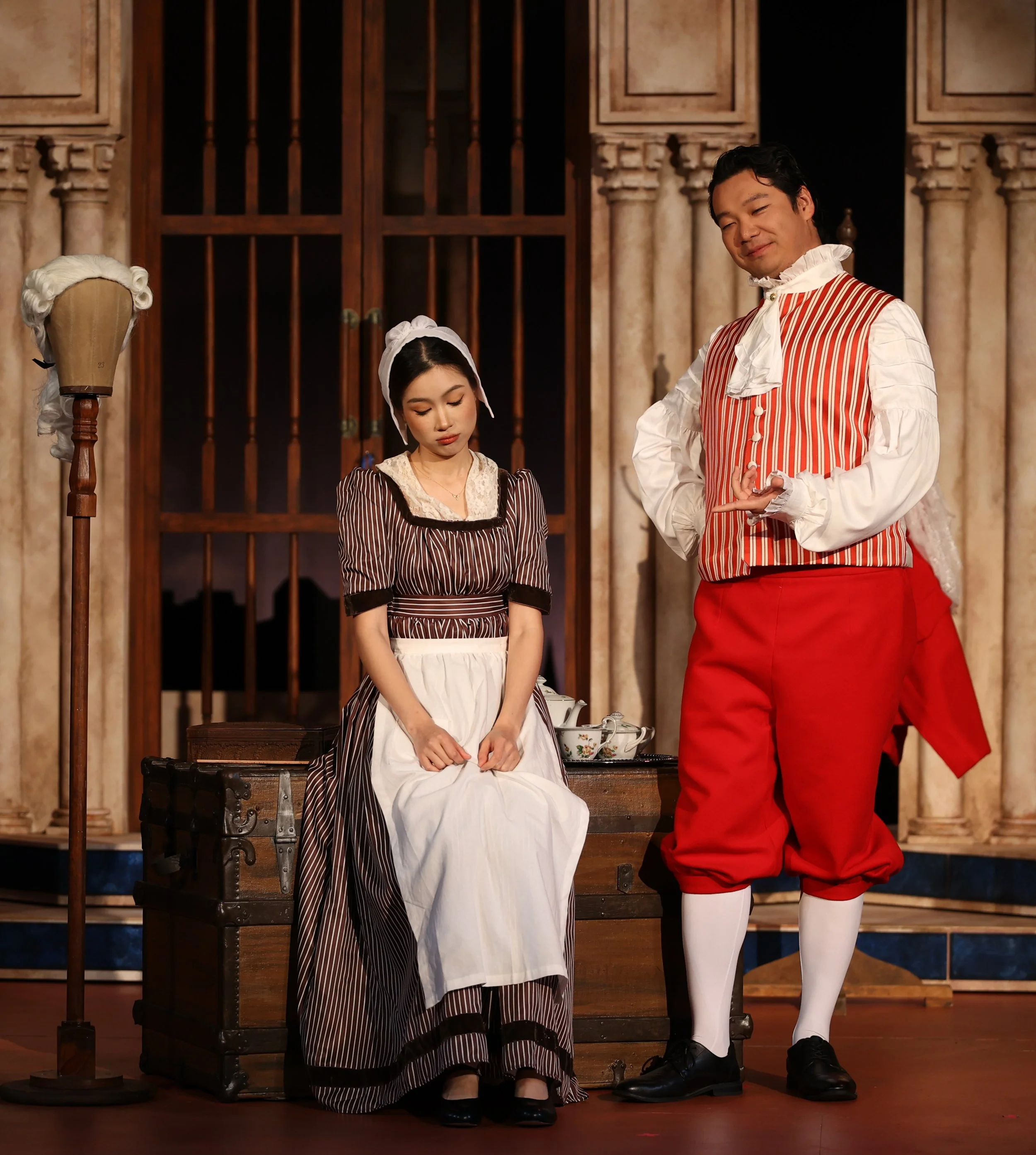 Two actors in historical costumes, one woman seated with her head bowed and a man standing beside her with a slight smile, set against a vintage wooden backdrop with columns and a barred window, evoking a period drama scene.