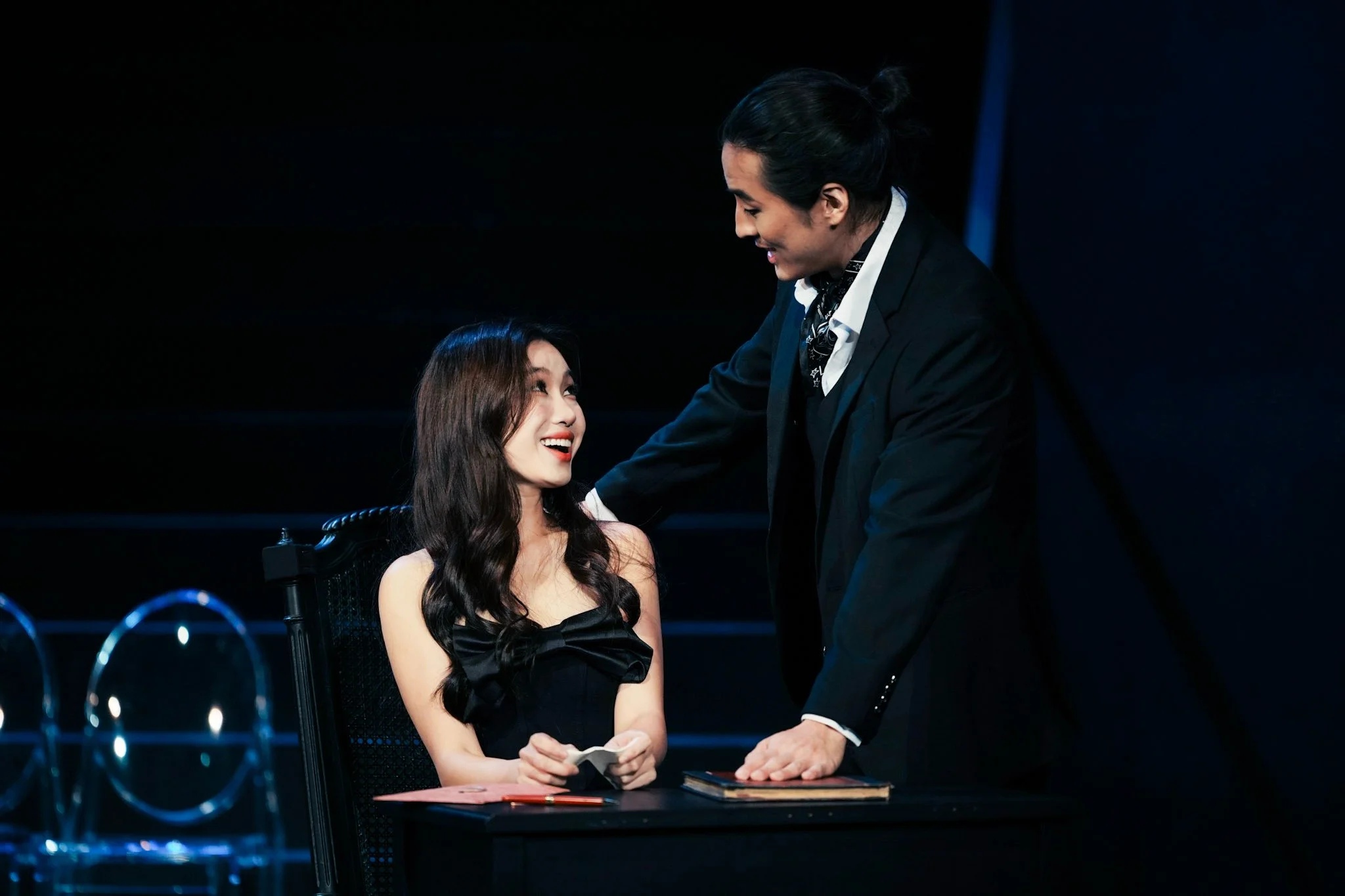 A man in a black suit and patterned scarf talking to a woman in a black dress with wavy hair, sitting at a table with a red folder, pens, and a smartphone, in a dark room with a black background.