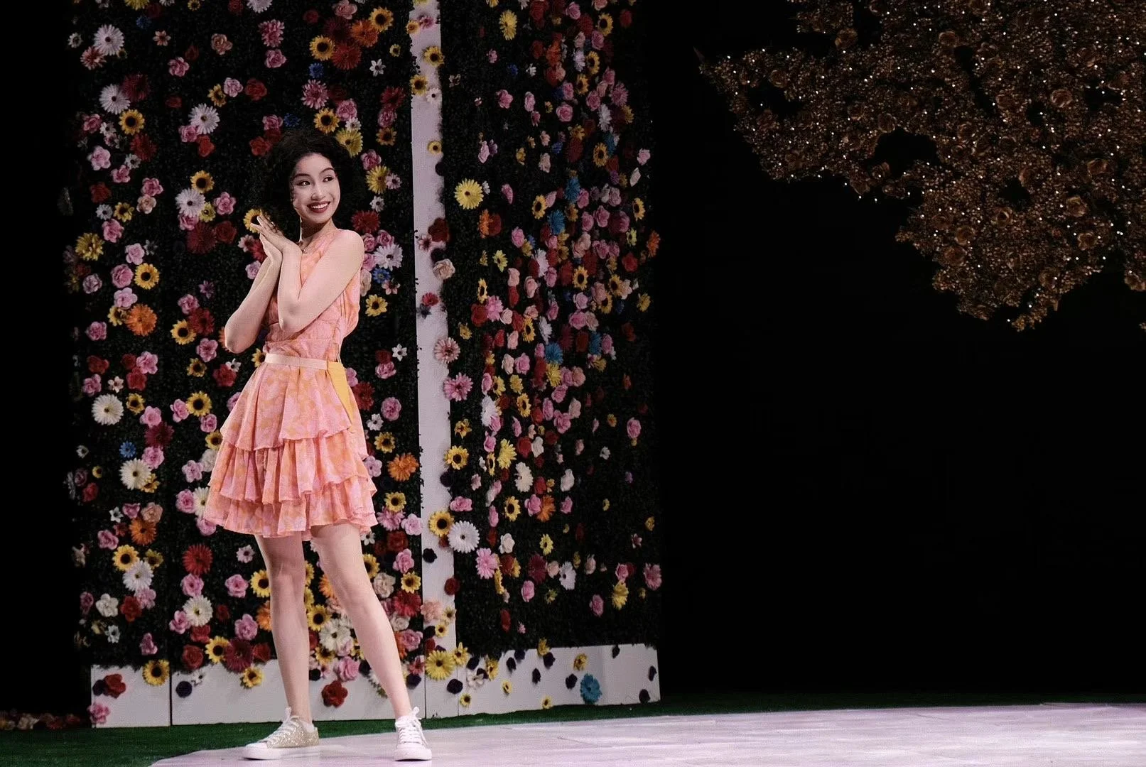 A young girl in a pink floral dress standing on a stage with a backdrop of colorful flowers and dark decorative elements.