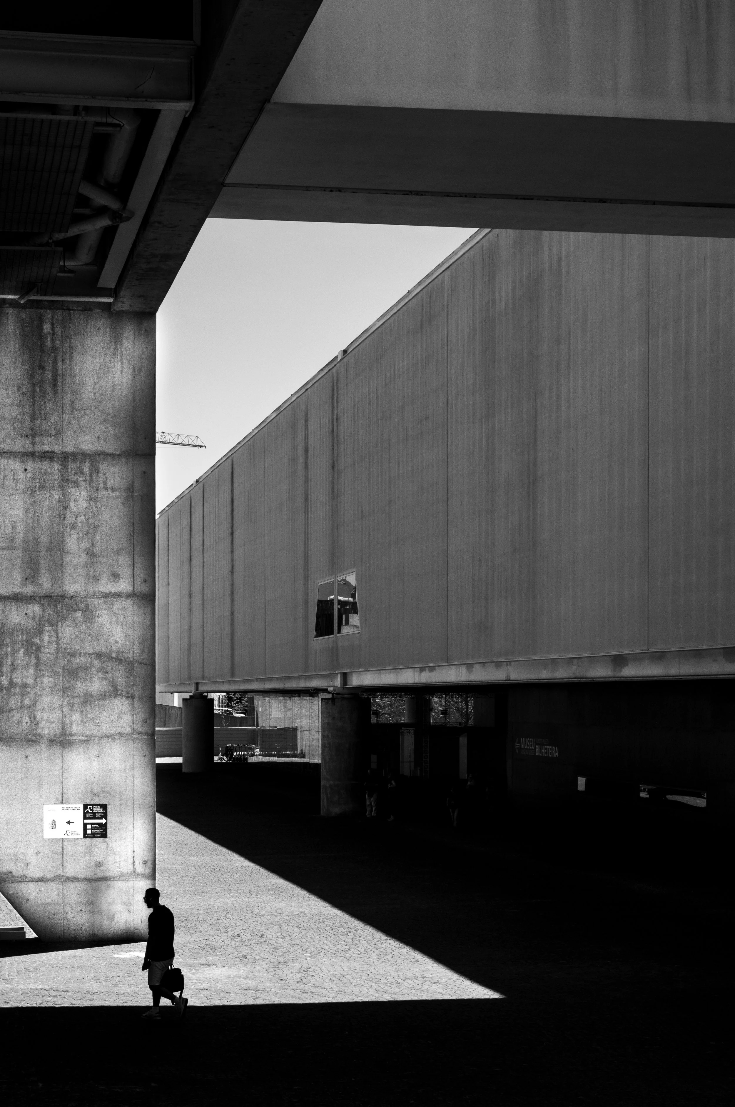 A black-and-white photo of a modern building with large concrete pillars and geometric shapes, featuring a few people walking underneath and around the structure.
