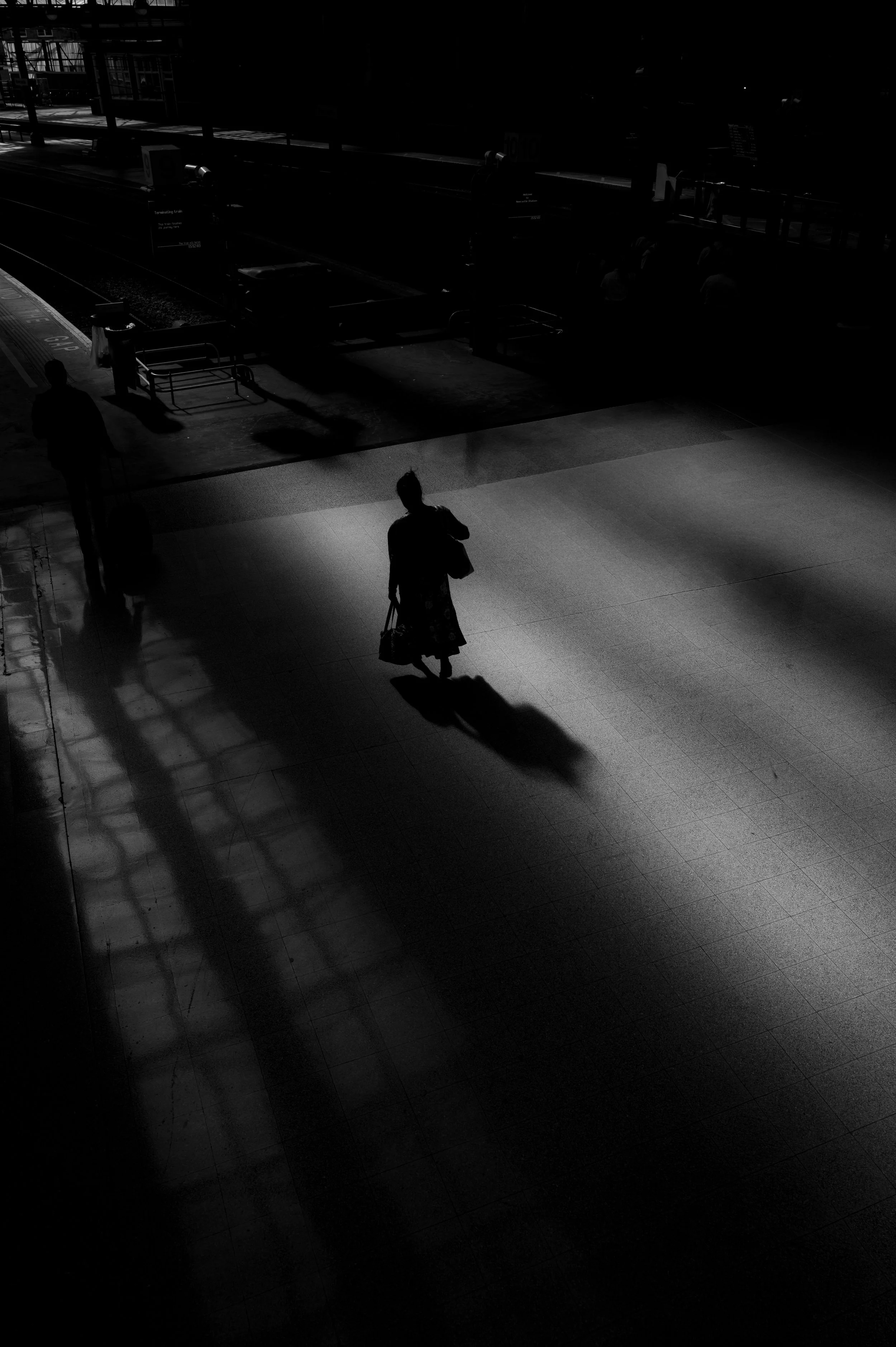 A dark, shadowy scene of a train station platform with a few silhouetted people, illuminated by sparse light, creating stark shadows on the ground.