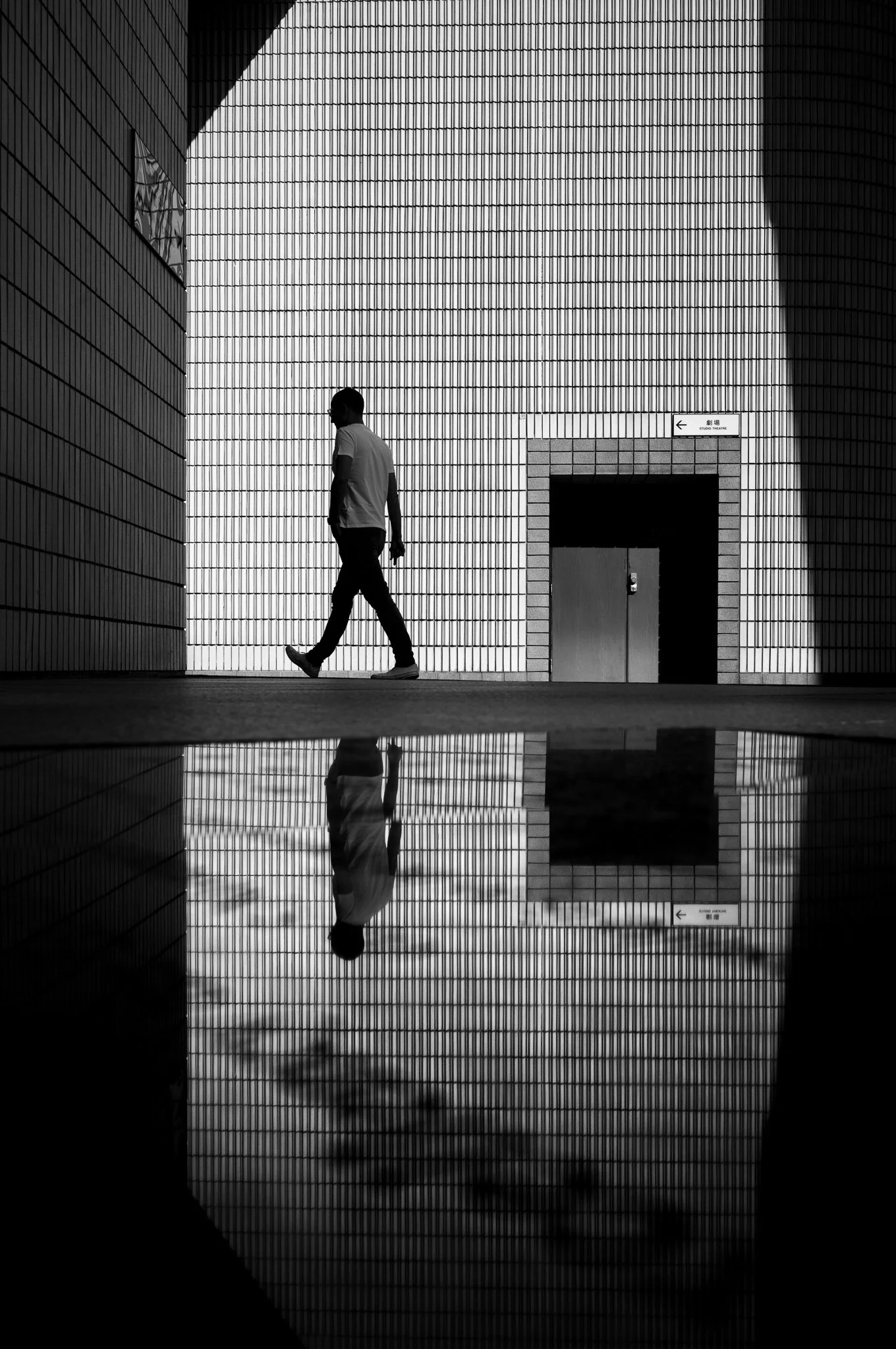 A person walking indoors near a large illuminated wall with horizontal tiles, with their reflection visible on the shiny floor.