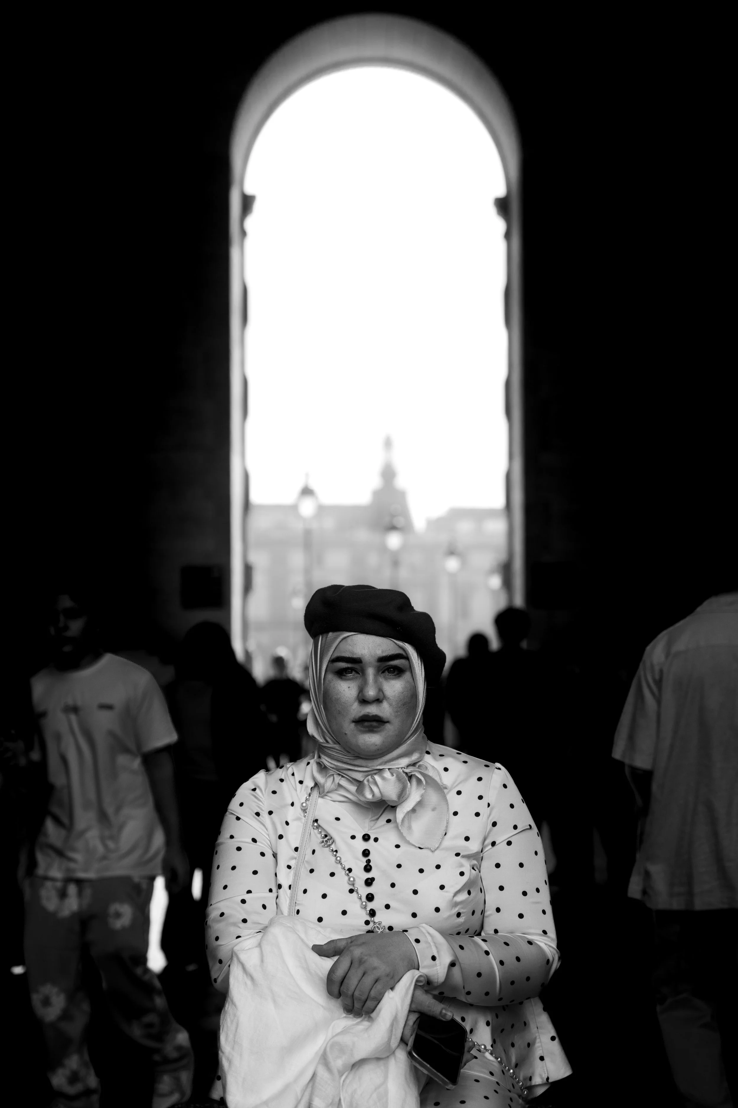 A woman wearing a polka dot shirt, headscarf, beret, and pearl necklace standing with arms crossed among a crowd, with an arched window in the background.