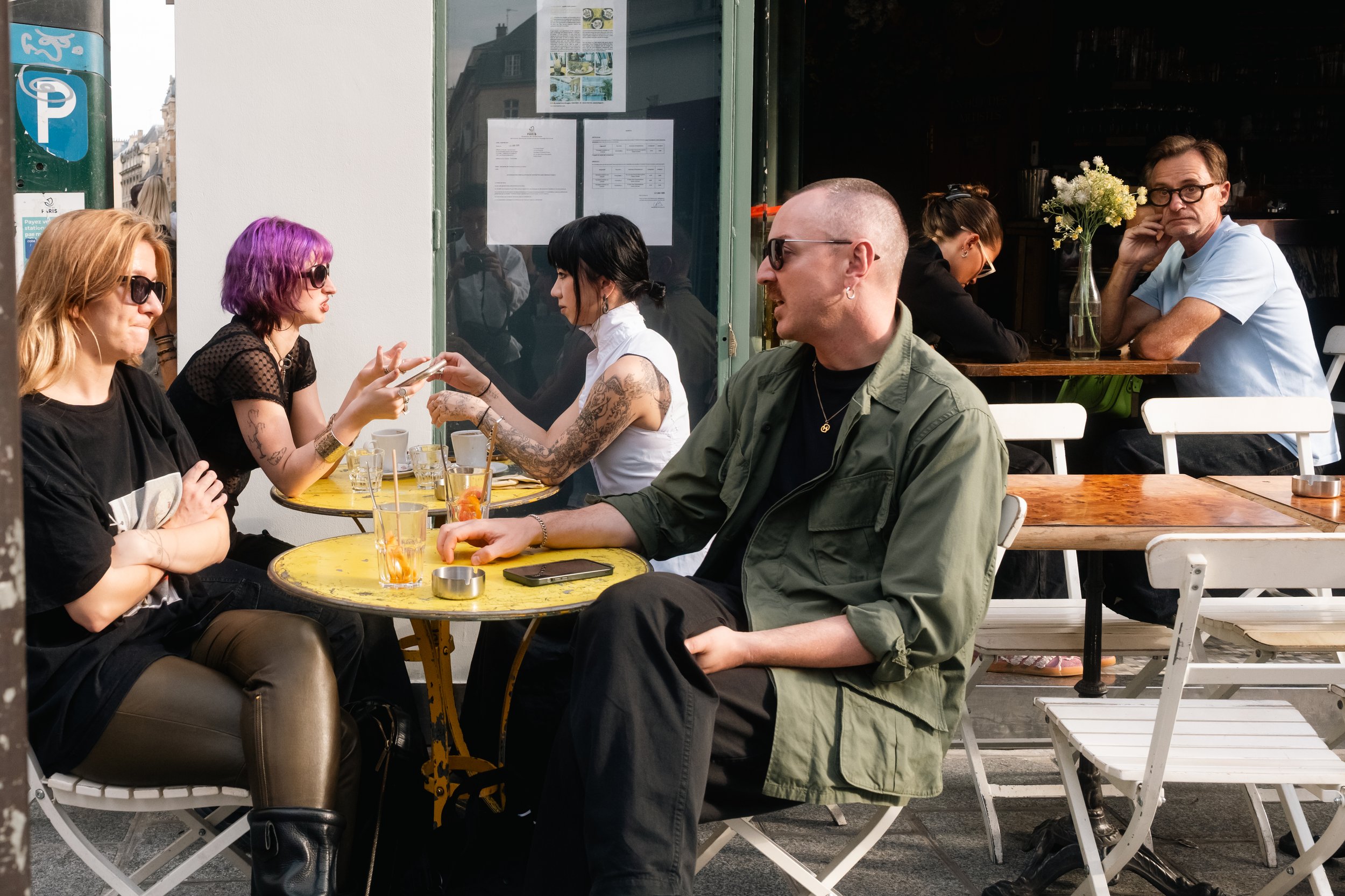 People sitting outside at a cafe, some are talking and using their phones, with drinks on the table and a man with glasses sitting alone at another table in the background.