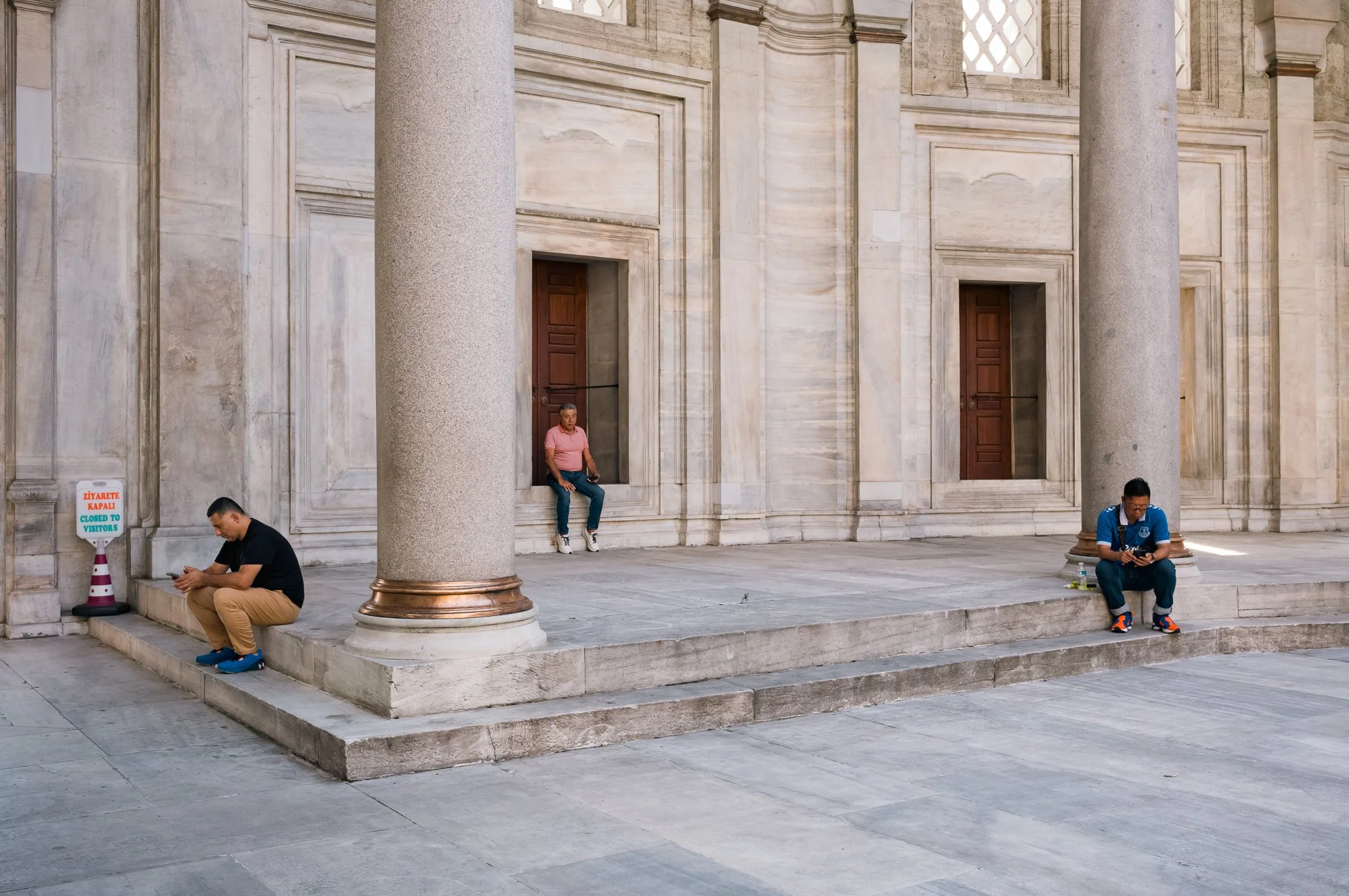 People sitting outside a large marble building with columns and wooden doors, some using their phones and one leaning against a column.