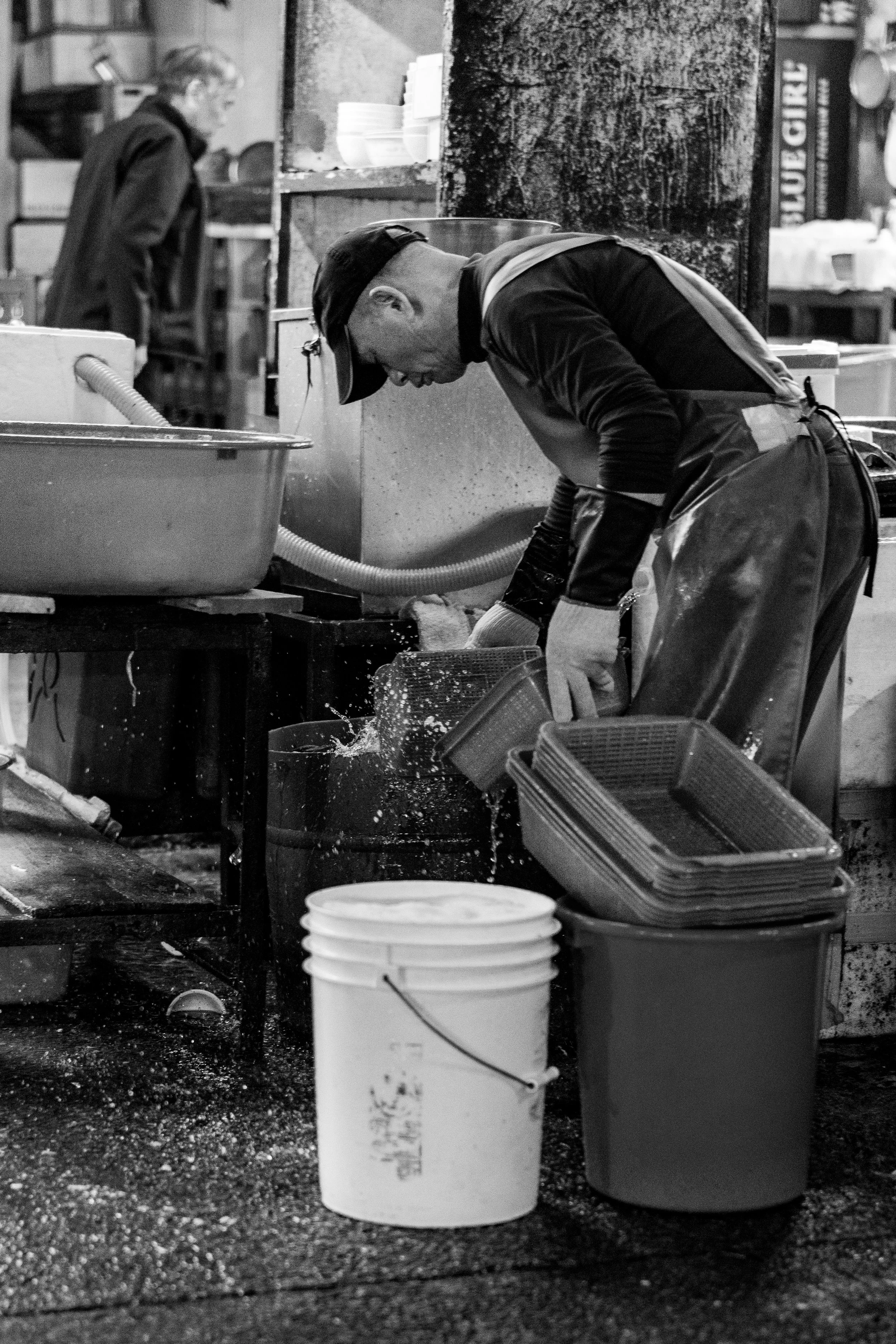 Two men working in a commercial kitchen, one washing dishes and the other in the background at another station, with buckets and trays around them.