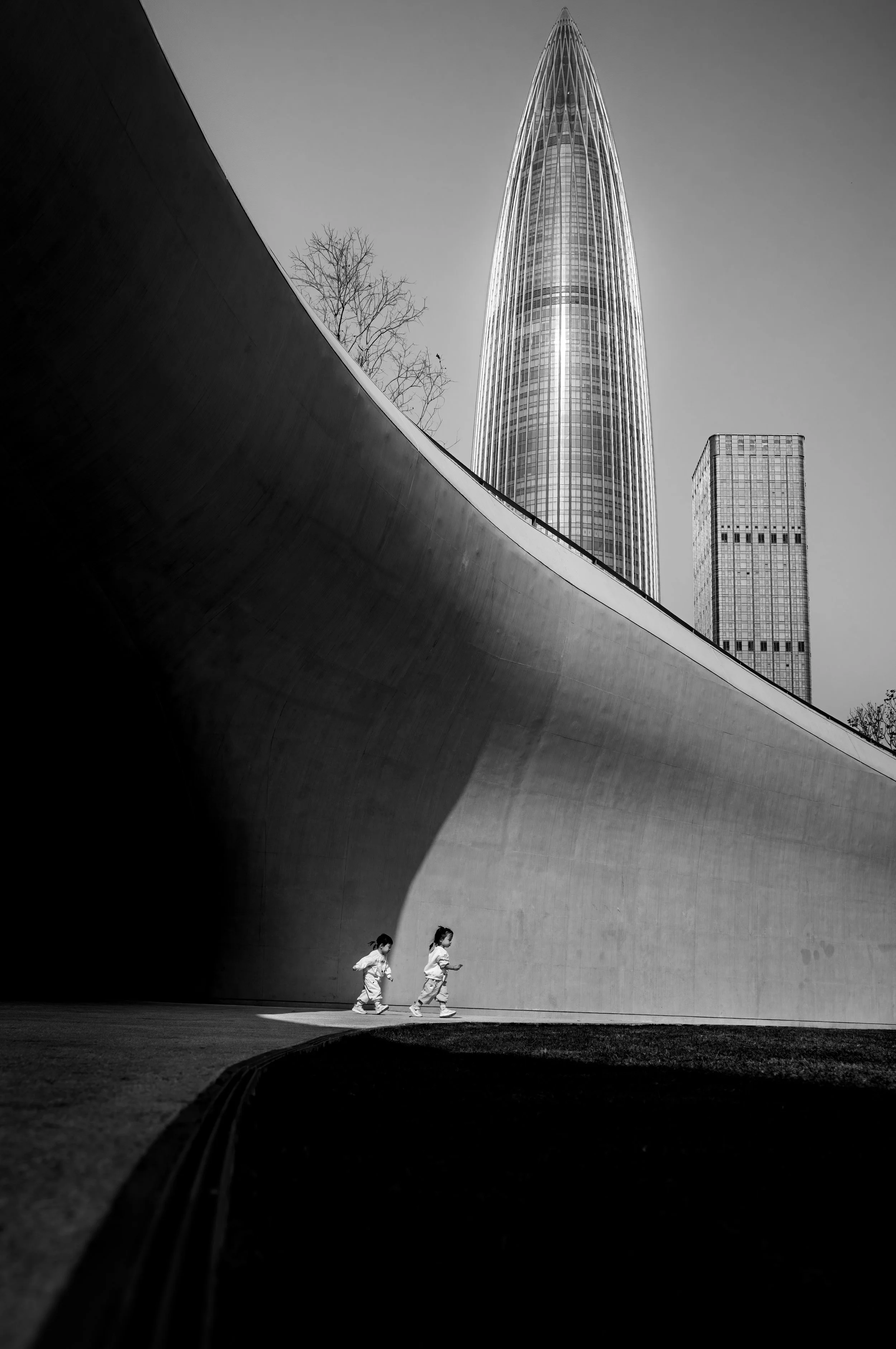 Black and white photograph of two children running in a futuristic urban setting. The background features towering skyscrapers with modern architecture.