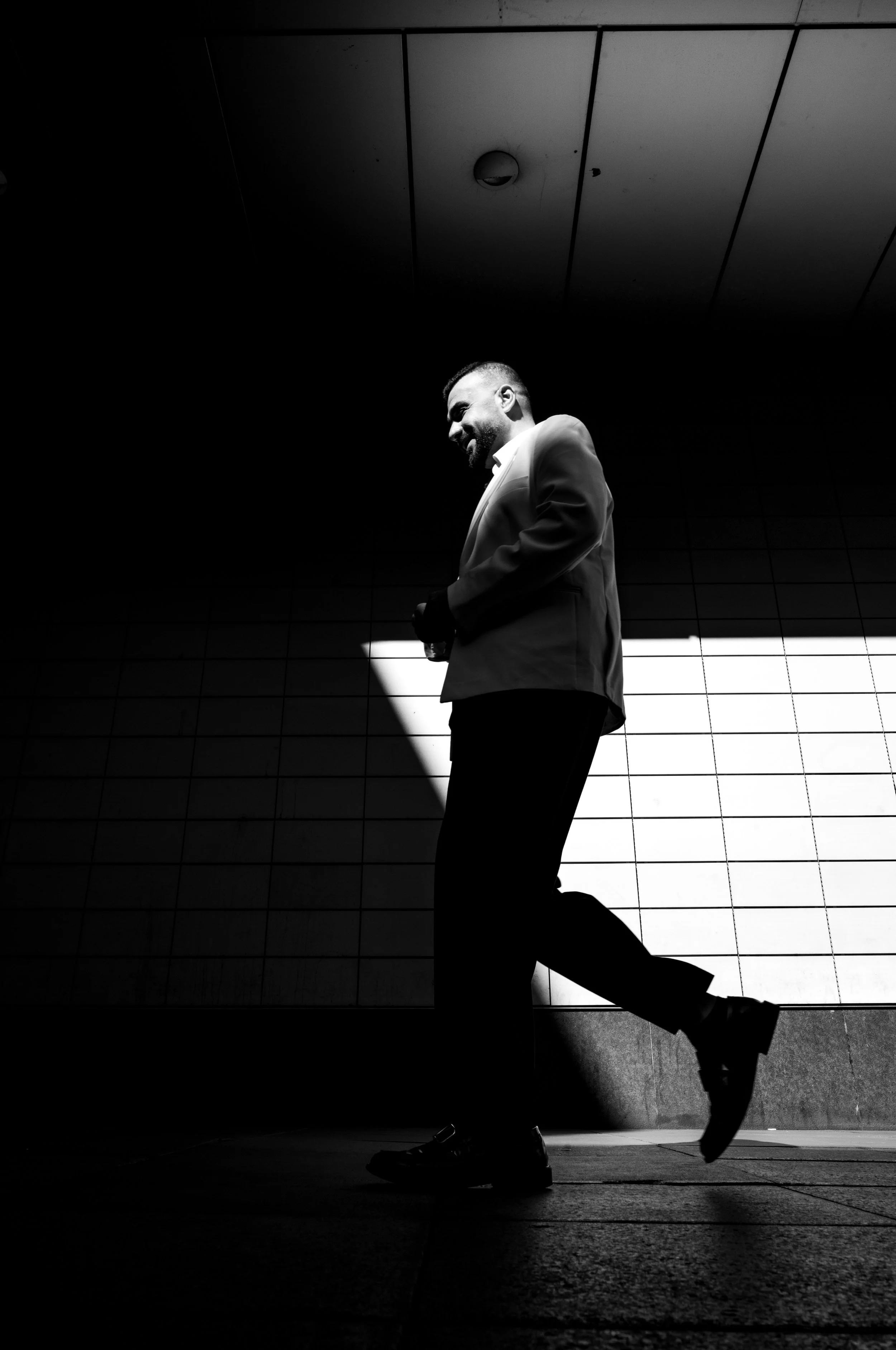 Black and white photo of a man in a suit walking in front of a tiled wall with a large bright light behind him, creating a silhouette effect.