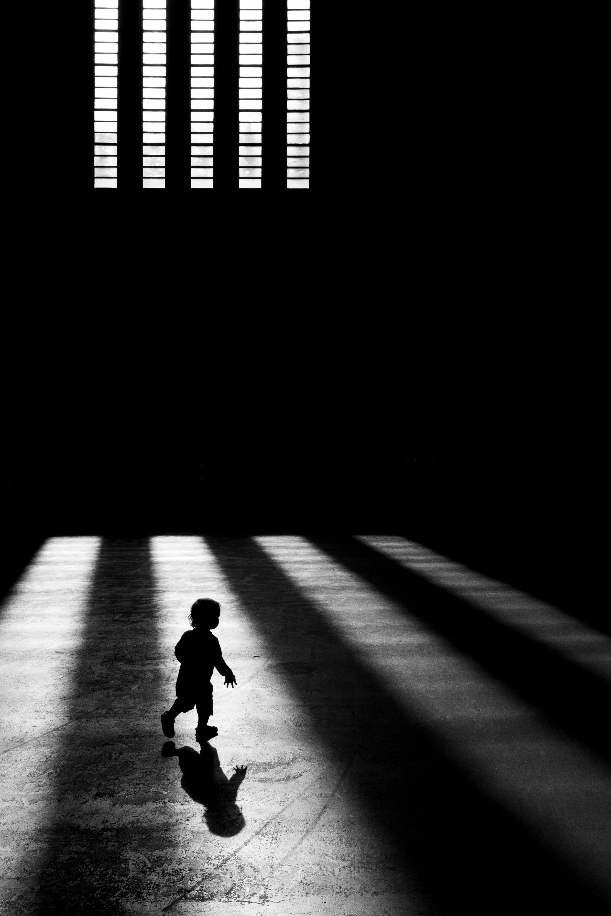 Silhouette of a young child standing in a dark room with sunlight coming through windows, casting shadows on the floor.