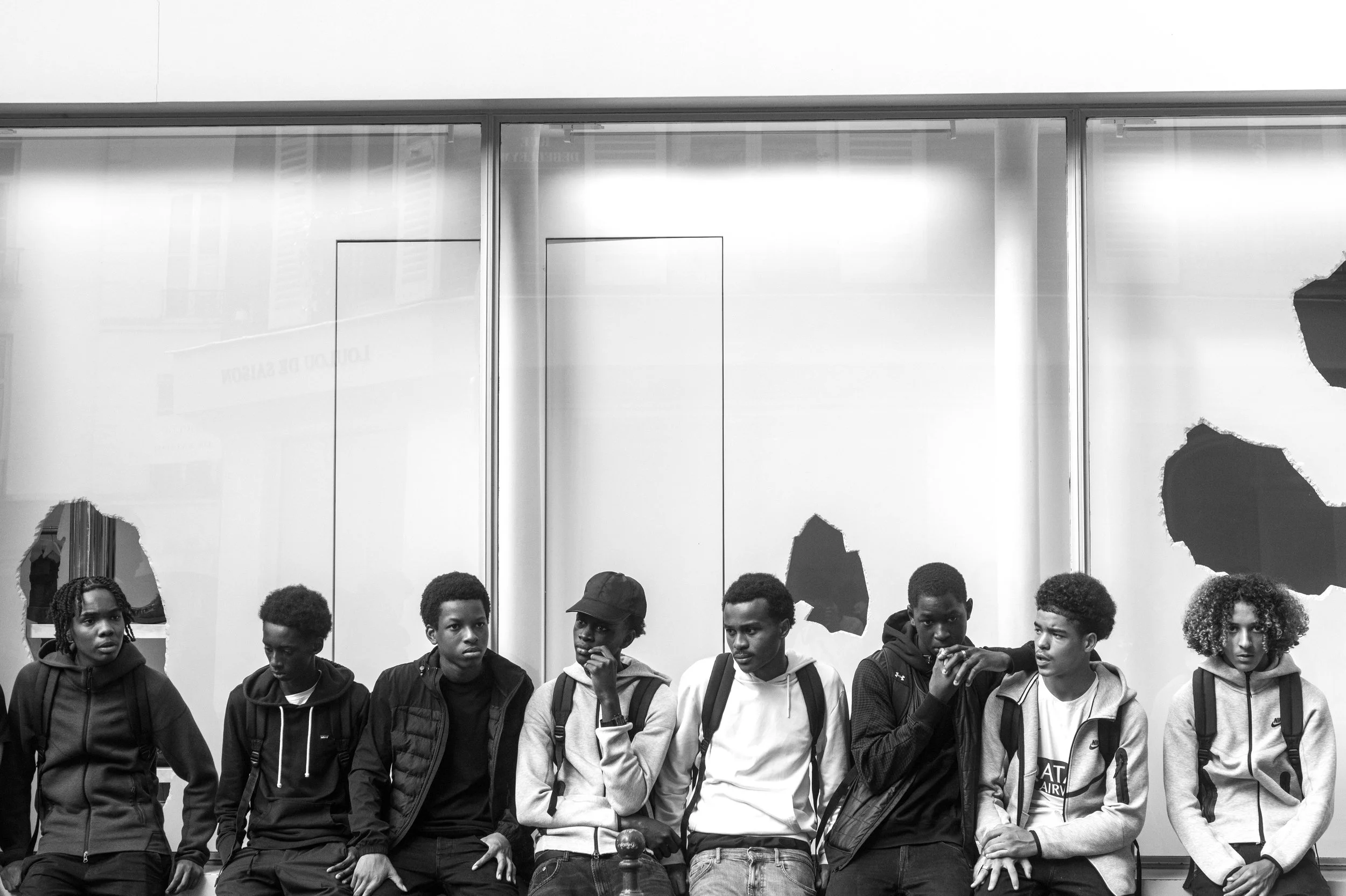 A group of young people of various ethnicities standing in front of a mirrored wall with torn sections, in what appears to be a public or communal space.