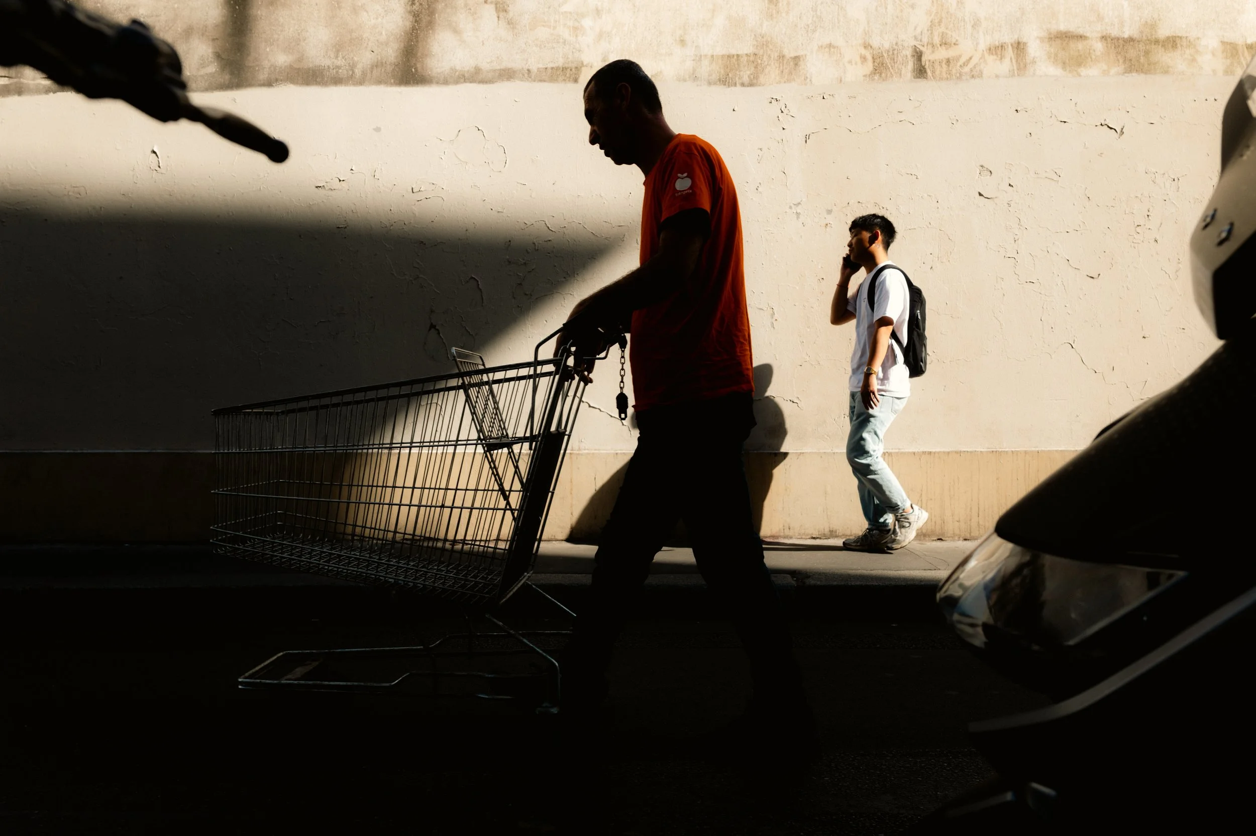 A man in an orange shirt and a man in a white shirt walking along a textured beige wall, with a shopping cart on the sidewalk, and part of a car visible in the lower right corner.