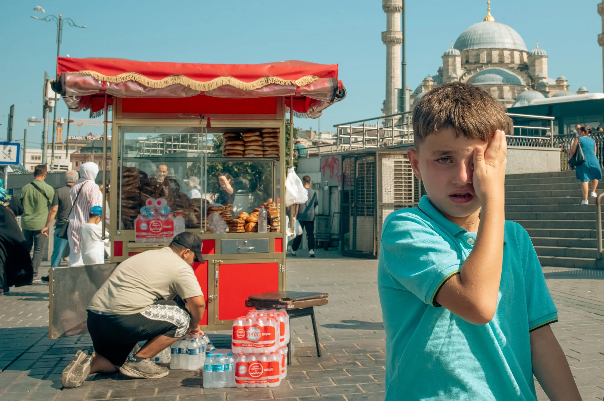 A young boy with brown hair wearing a blue shirt with black trim, covering one eye with his hand, standing in an outdoor area with a food stand selling baked goods, surrounded by people, and historic buildings in the background, including a domed mosque.