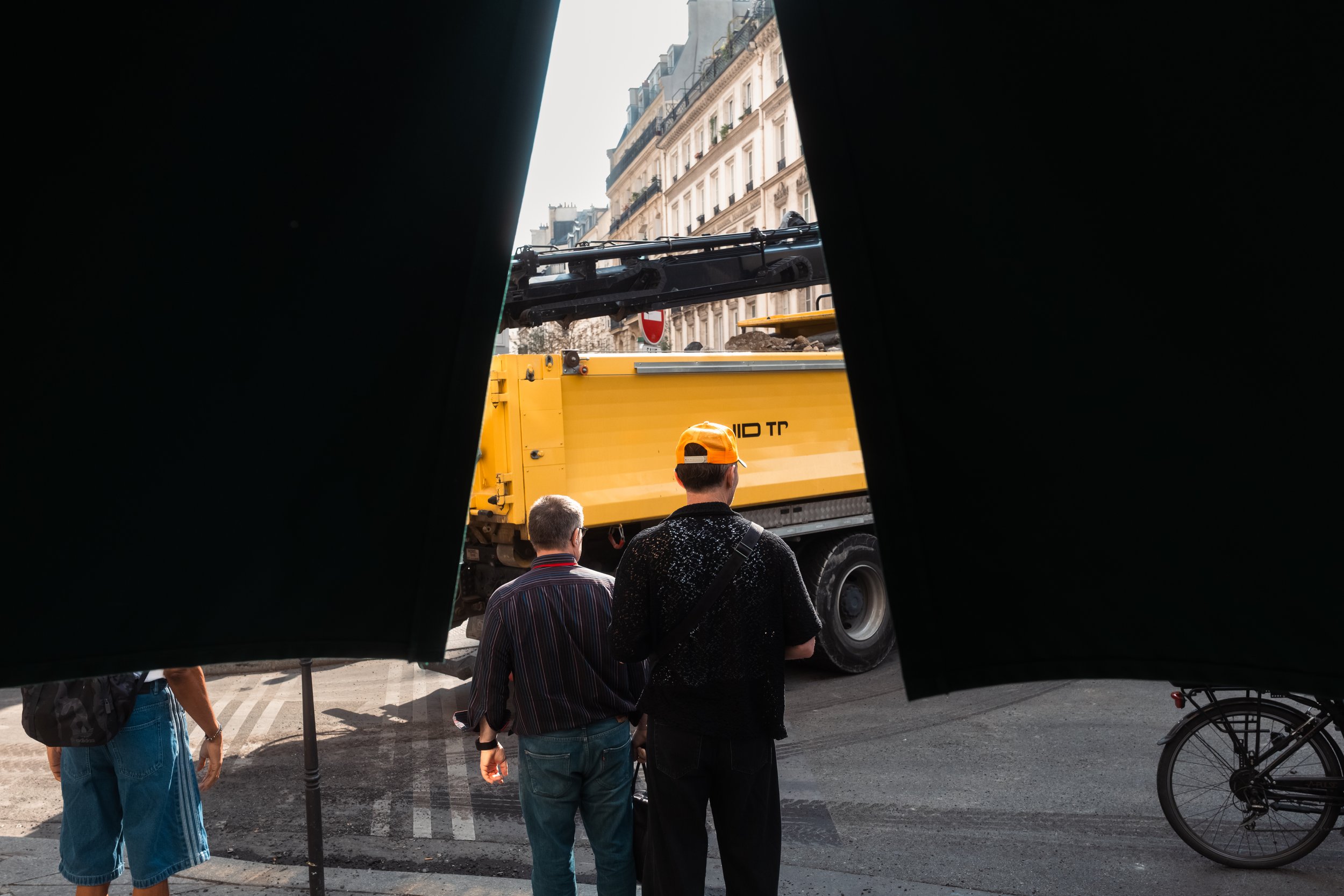 View of a city street through a narrow opening, showing people standing near a yellow construction truck. Buildings with classic European architecture are visible in the background.