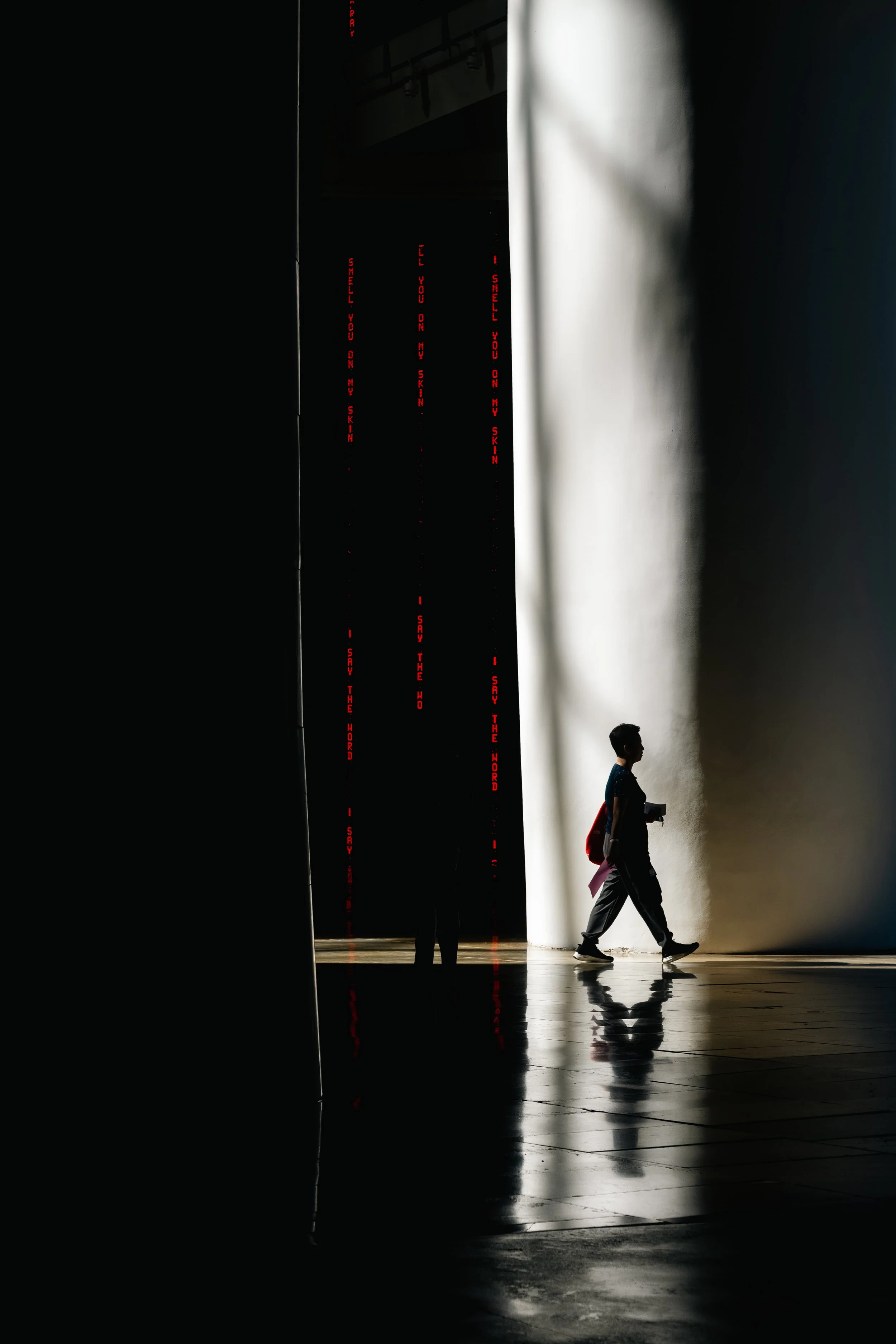 A person walking past a white wall illuminated with natural light, with a shadow cast on the wall. There are red vertical digital signs with text that reads 'I SAY THE WORD,' 'SMELL YOU ON MY SKIN,' 'I WILL YOU ON MY SKIN,' and 'I SAY THE WORD.' The 