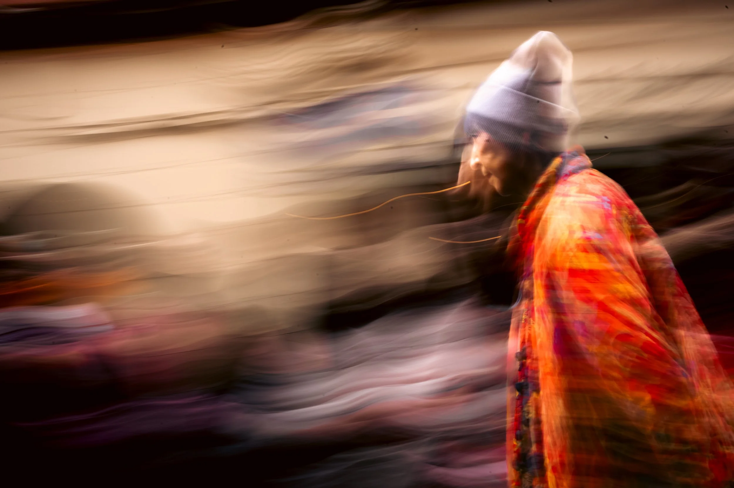 A person wearing a gray beanie and a colorful orange and red patterned jacket walking outdoors at night, with motion blur in the background.