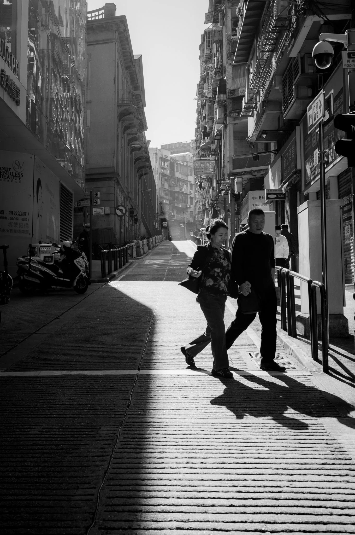 Street scene in an urban area with people walking and buildings on either side. Shadows cast on the sidewalk, with some signs and a motor scooter parked.