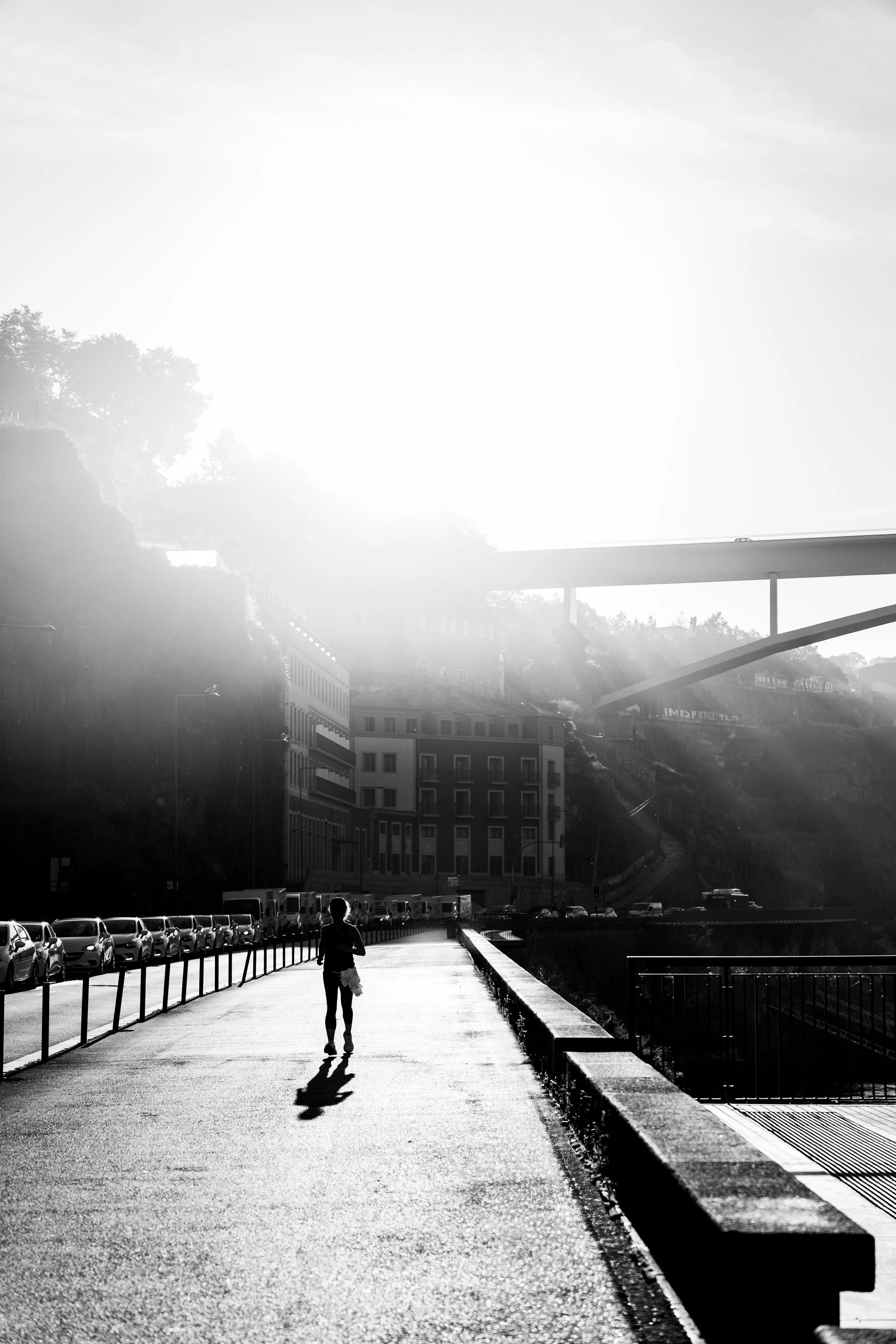 Person walking on a paved urban sidewalk with parked cars on the left, buildings on the right, and a bridge overhead in the background, with sunlight creating a high contrast scene.