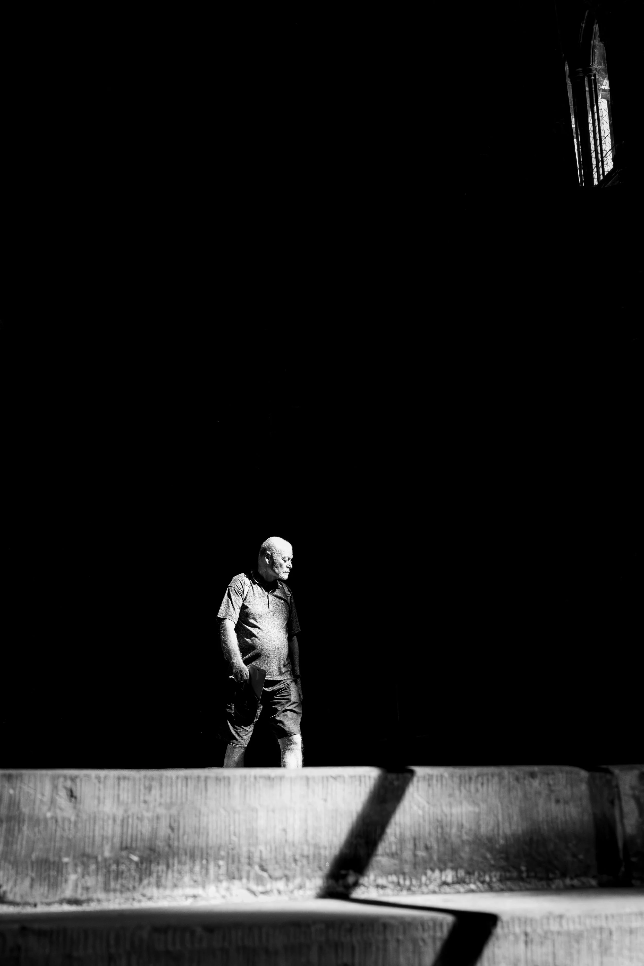 A man walking along a ledge under a large dark space with a window in the top right corner, captured in black and white.