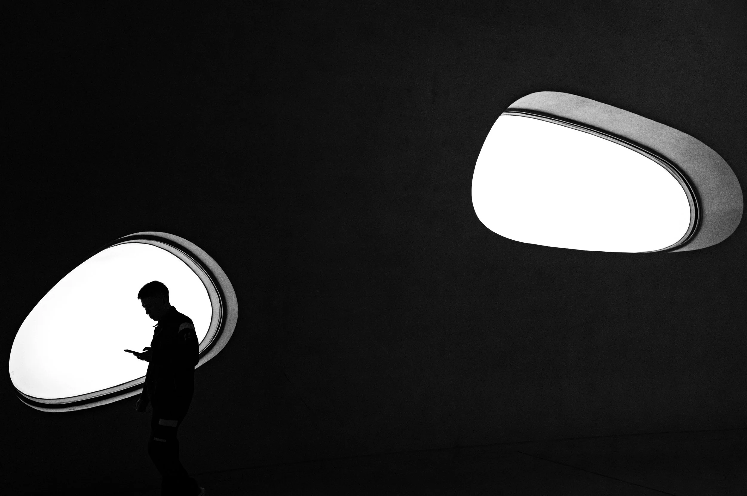 A person walking while looking at a mobile device in front of two illuminated, circular architectural openings with a dark background.