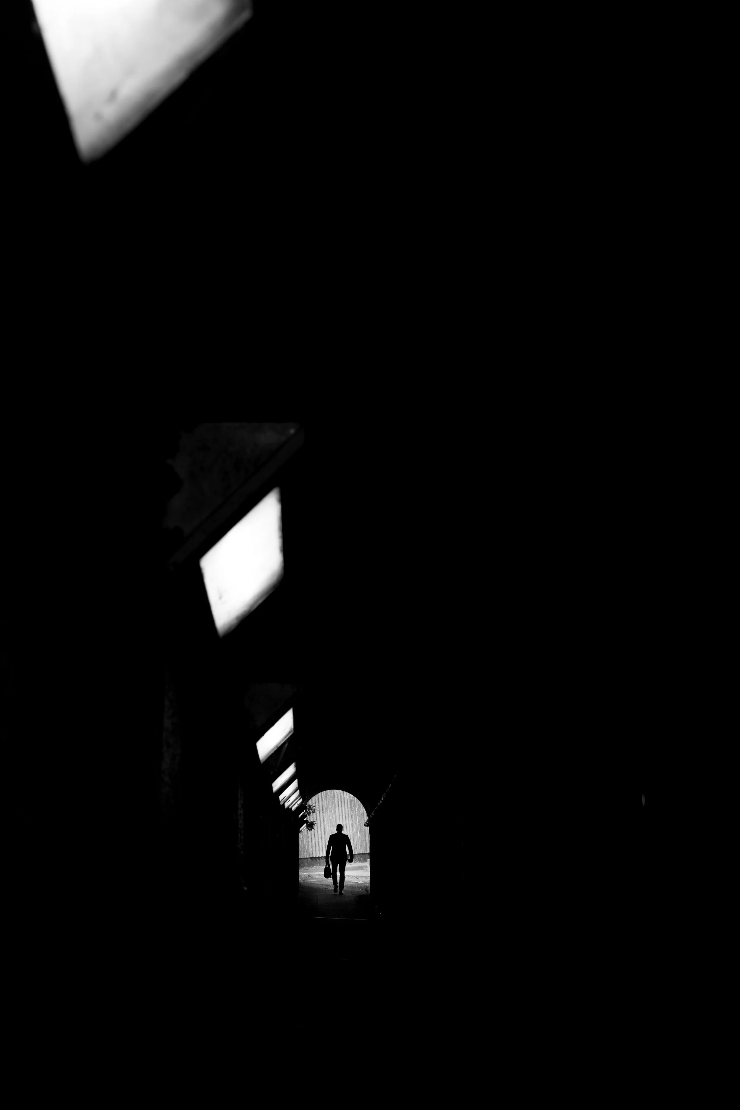 A silhouette of a person walking through a tunnel with light at the end, illuminated sequentially by rectangular light fixtures on the ceiling.