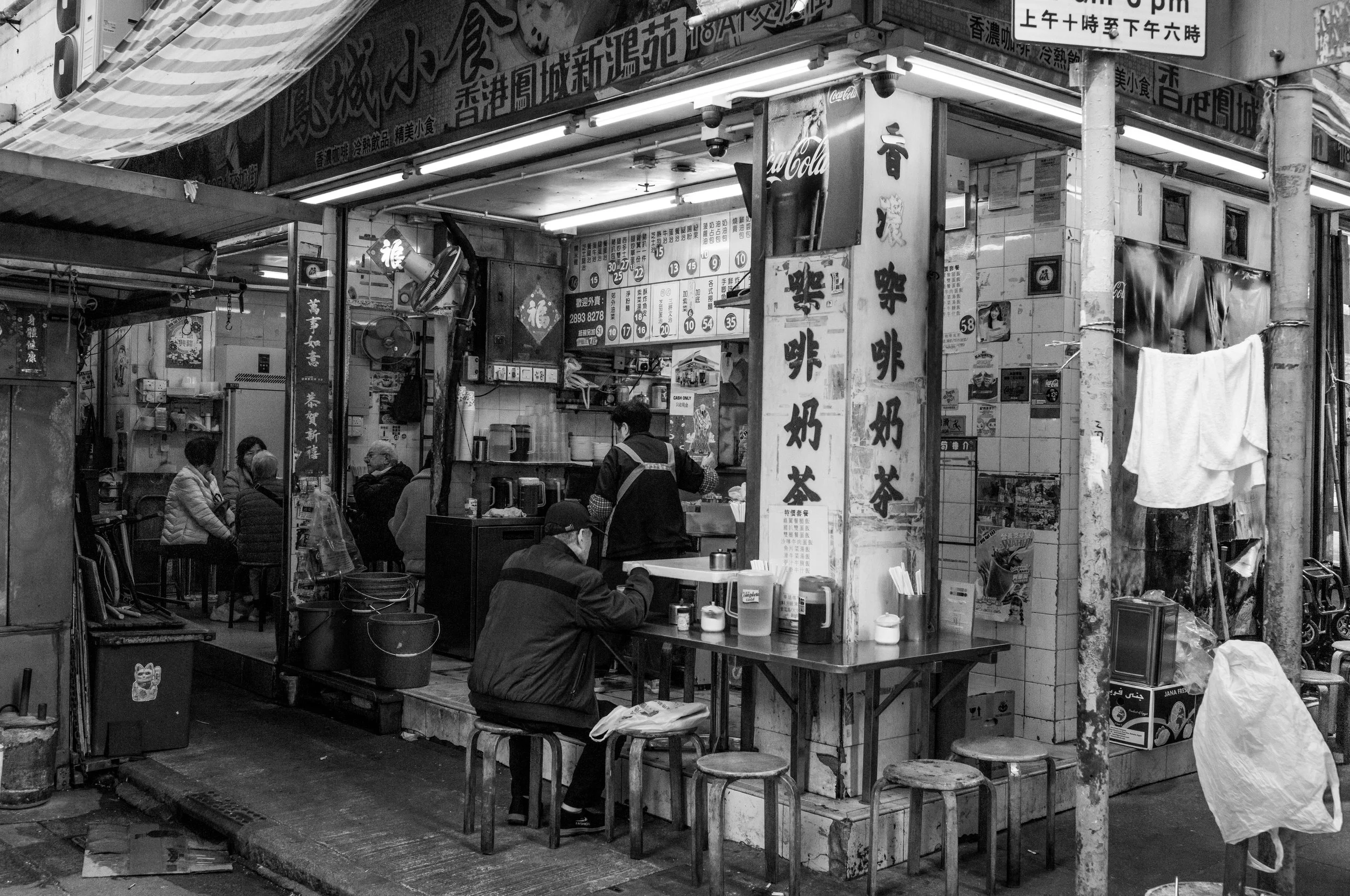 Street-side eatery with customers sitting at tables, menu signage in Chinese, and various food service items.