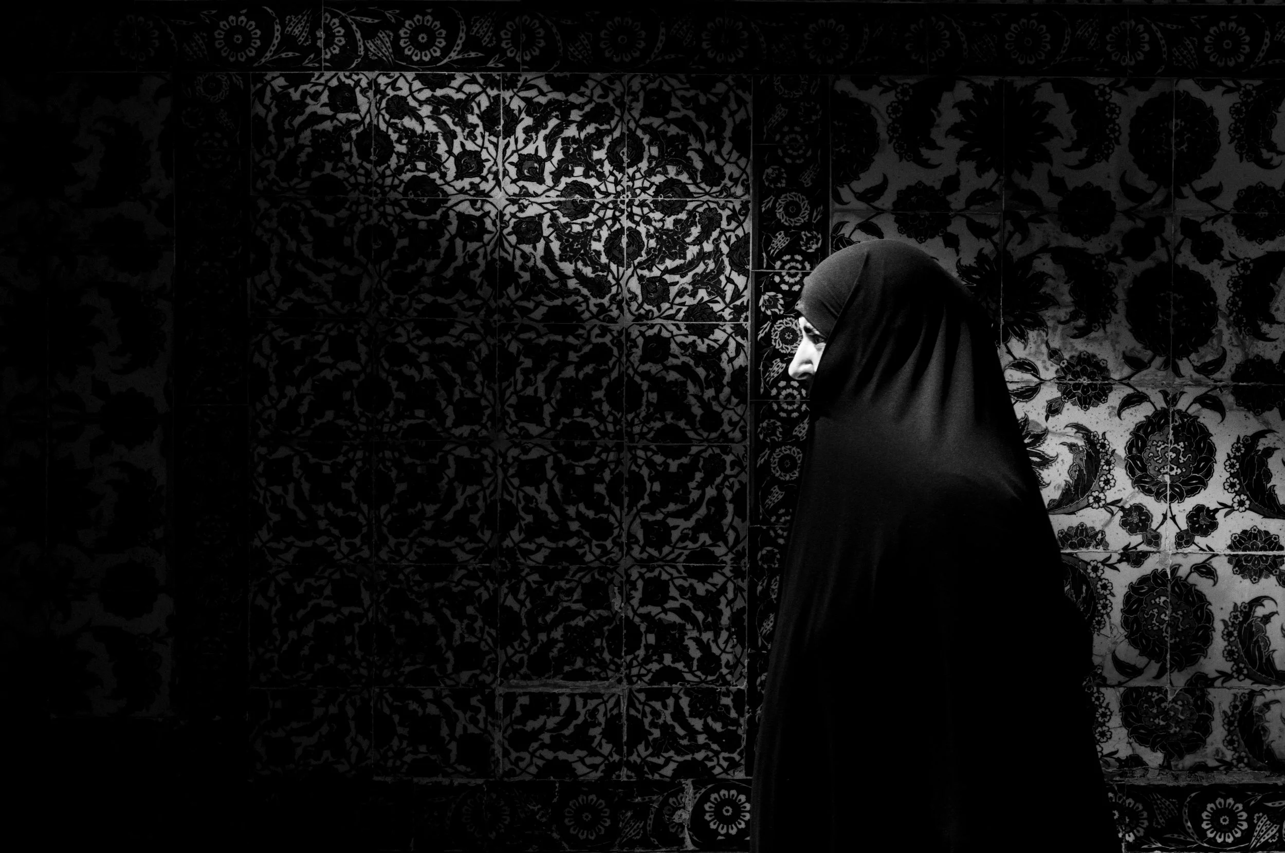 A woman wearing a headscarf walking past decorative tiles with floral and geometric patterns.
