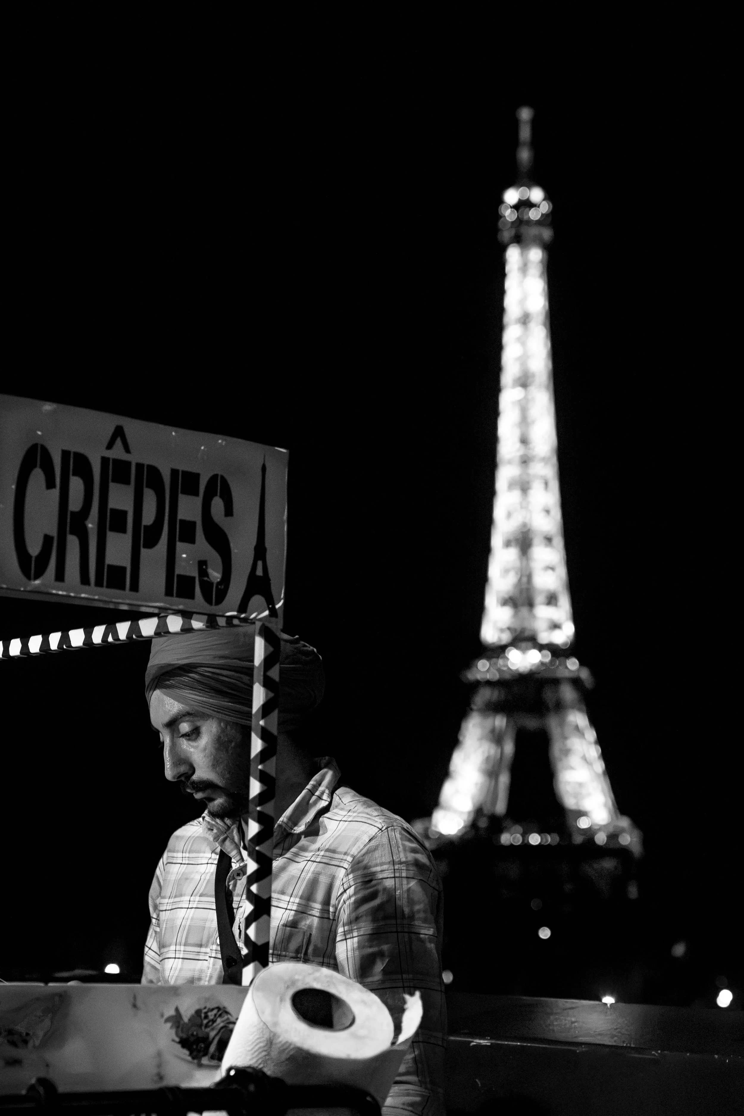 Night scene with a man at a food cart selling crepes, with the Eiffel Tower illuminated in the background.