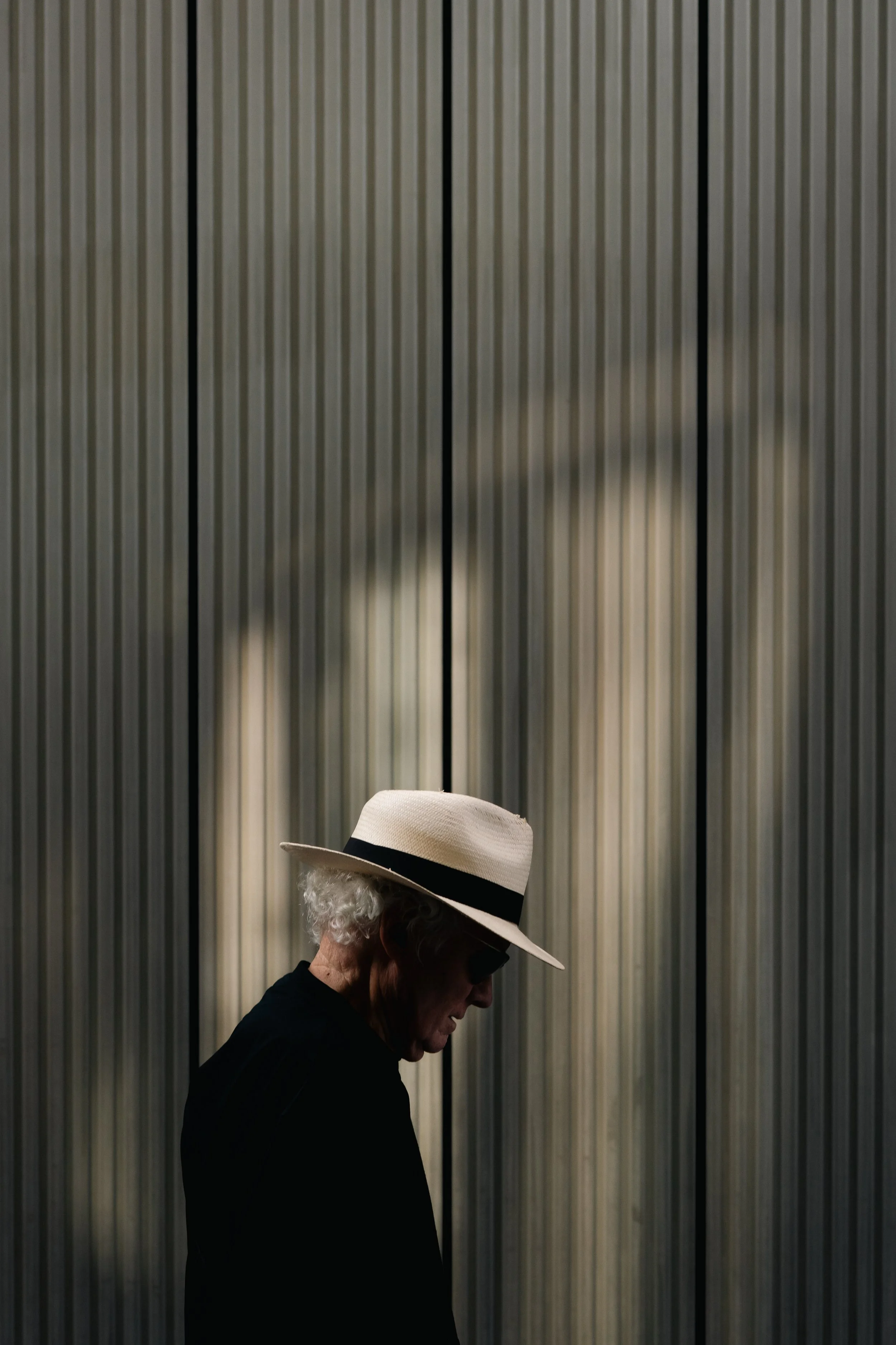 A woman with curly gray hair wearing a white hat with a black band and a black top, standing in front of a textured wall with vertical lines.