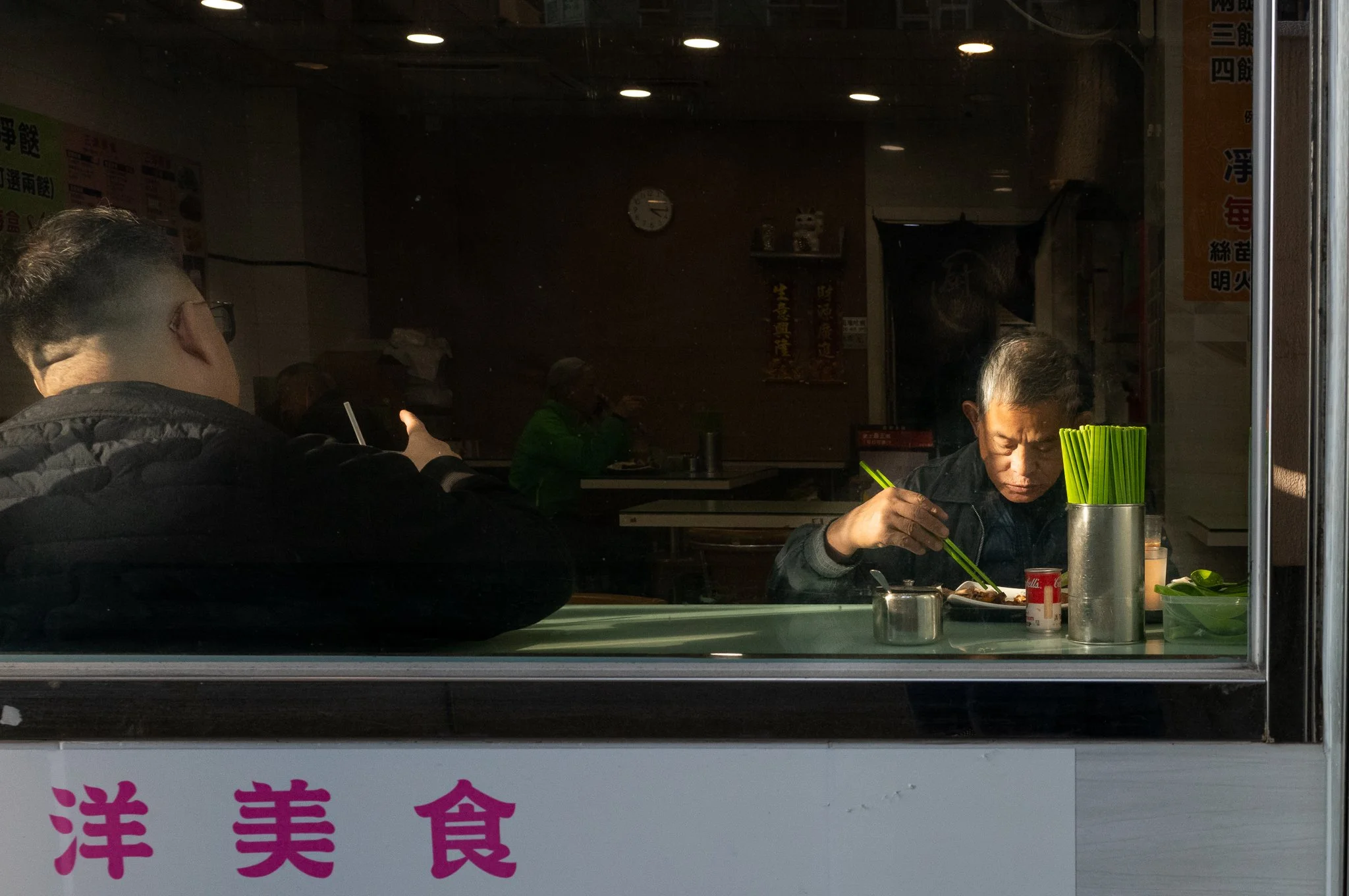 Inside a restaurant with dim lighting, two men are sitting at a table. One man, seen from behind, is using a straw. The other, on the right, is eating with chopsticks. Green napkins are in a cup, and a small can of Campbell's soup is on the table. Th