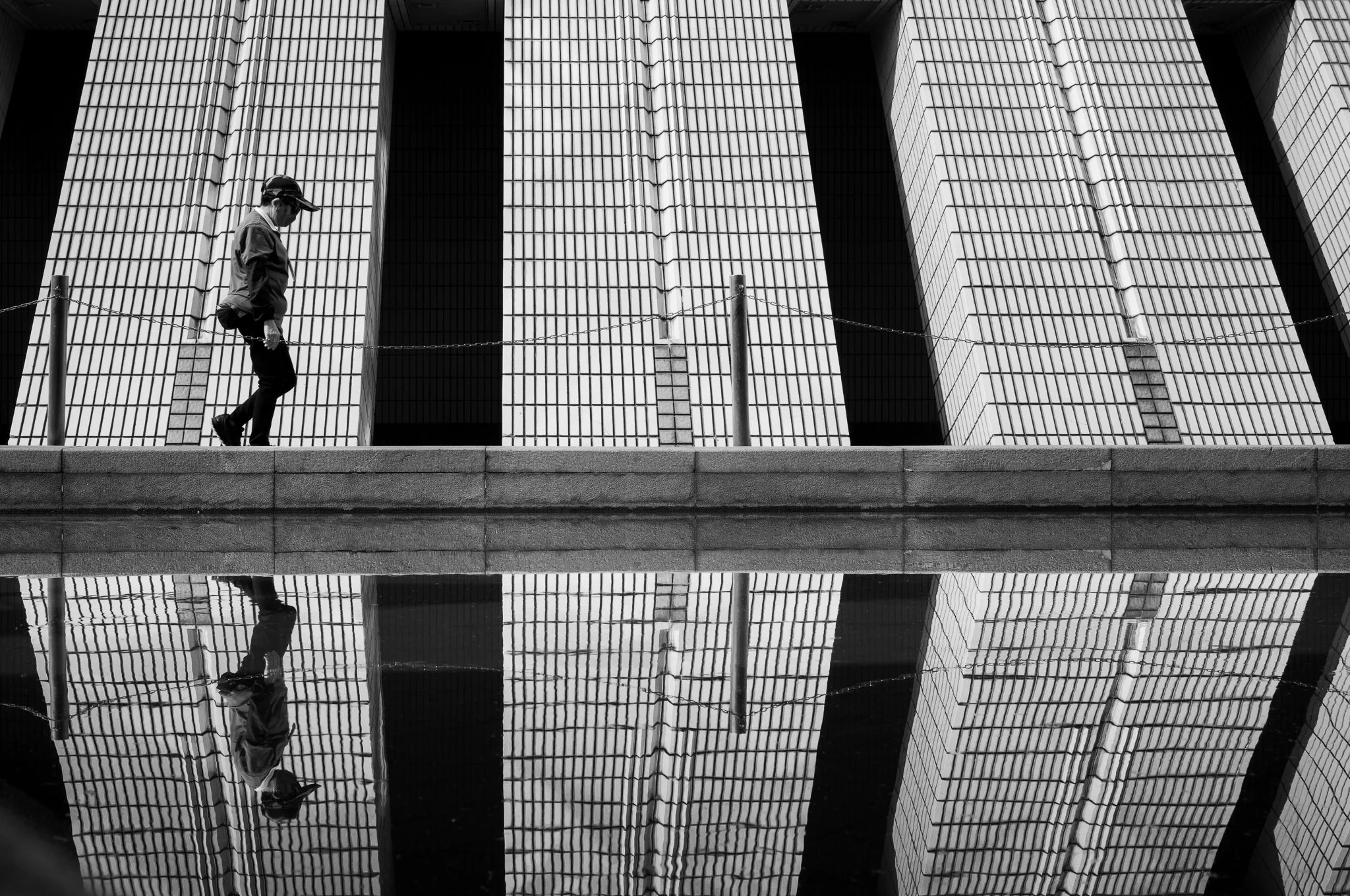 A man walking along a bridge or walkway in front of a tall modern building with a geometric facade, with a reflection of the building and the man in the water below.
