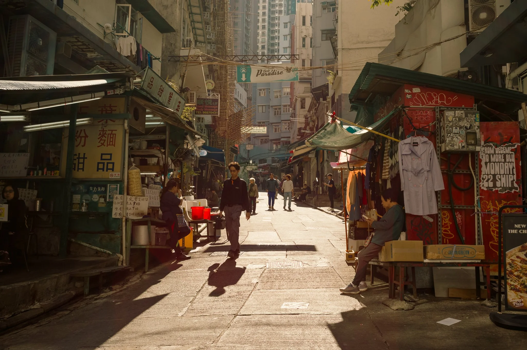 A busy street scene in an urban market area with small shops, stalls, and people walking and shopping. Tall buildings are visible in the background, with signs in Chinese and English.