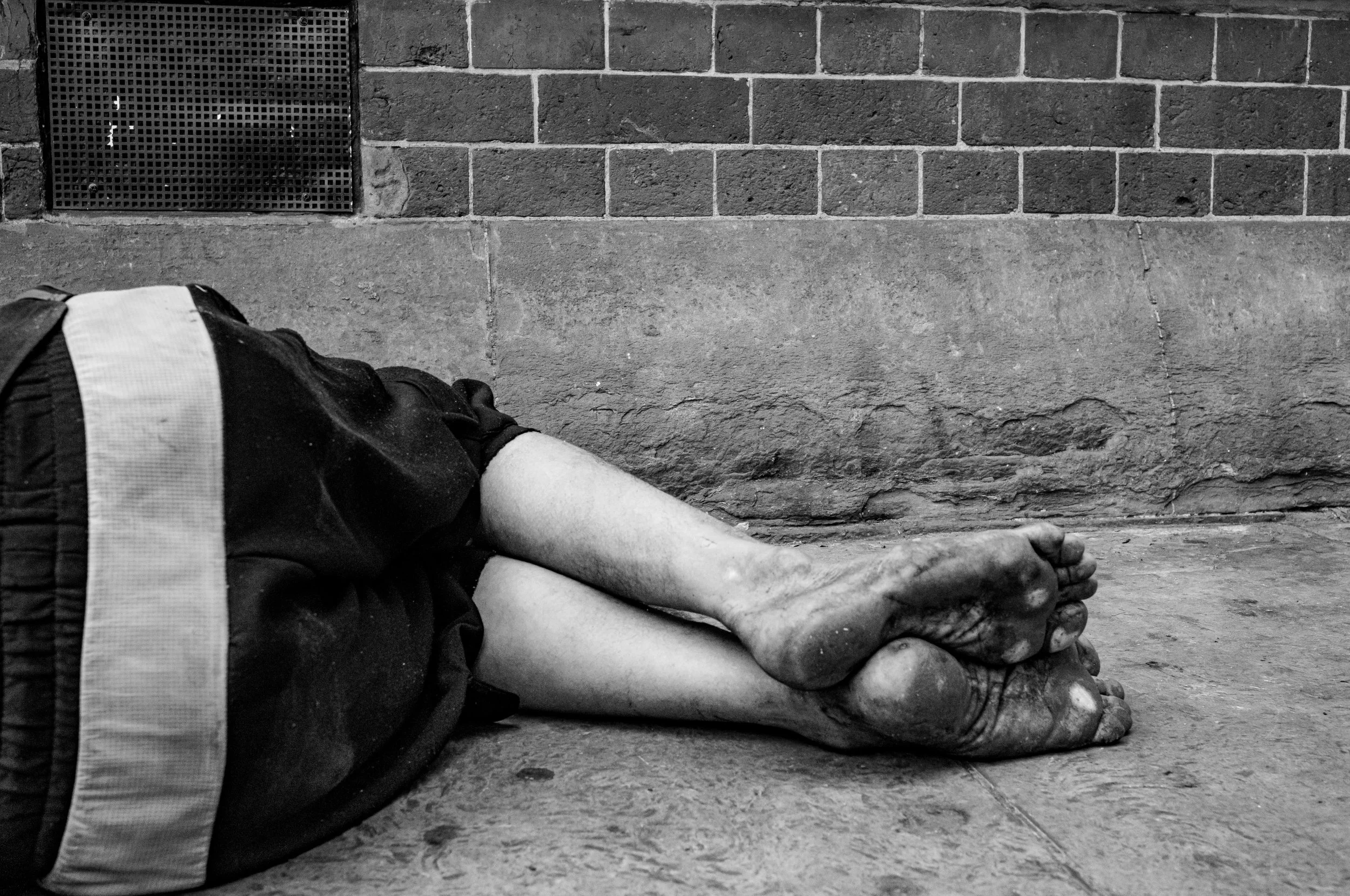A person lying on the ground with arms crossed and legs extended, wearing dark clothing with reflective stripes. The concrete floor and brick wall are visible, along with a metal grate in the wall.