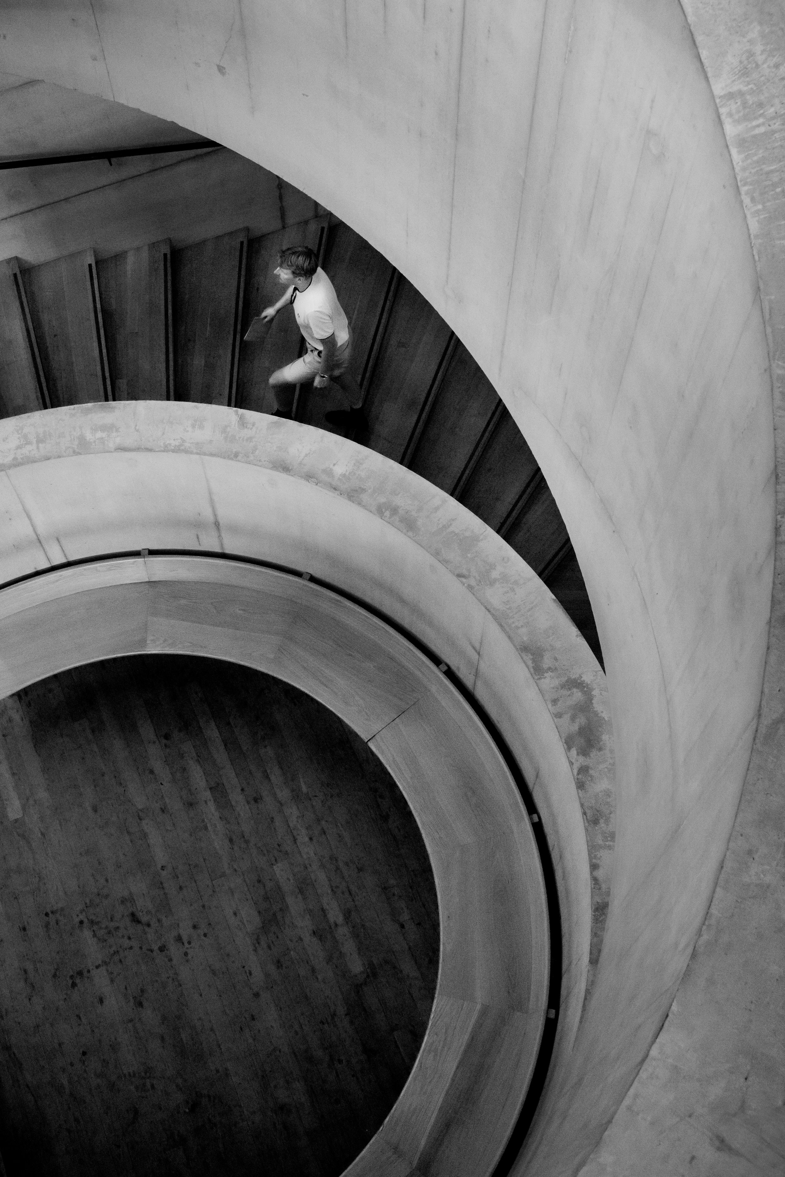 A man walking down a spiral staircase with concrete steps and wood-paneled walls, viewed from above.