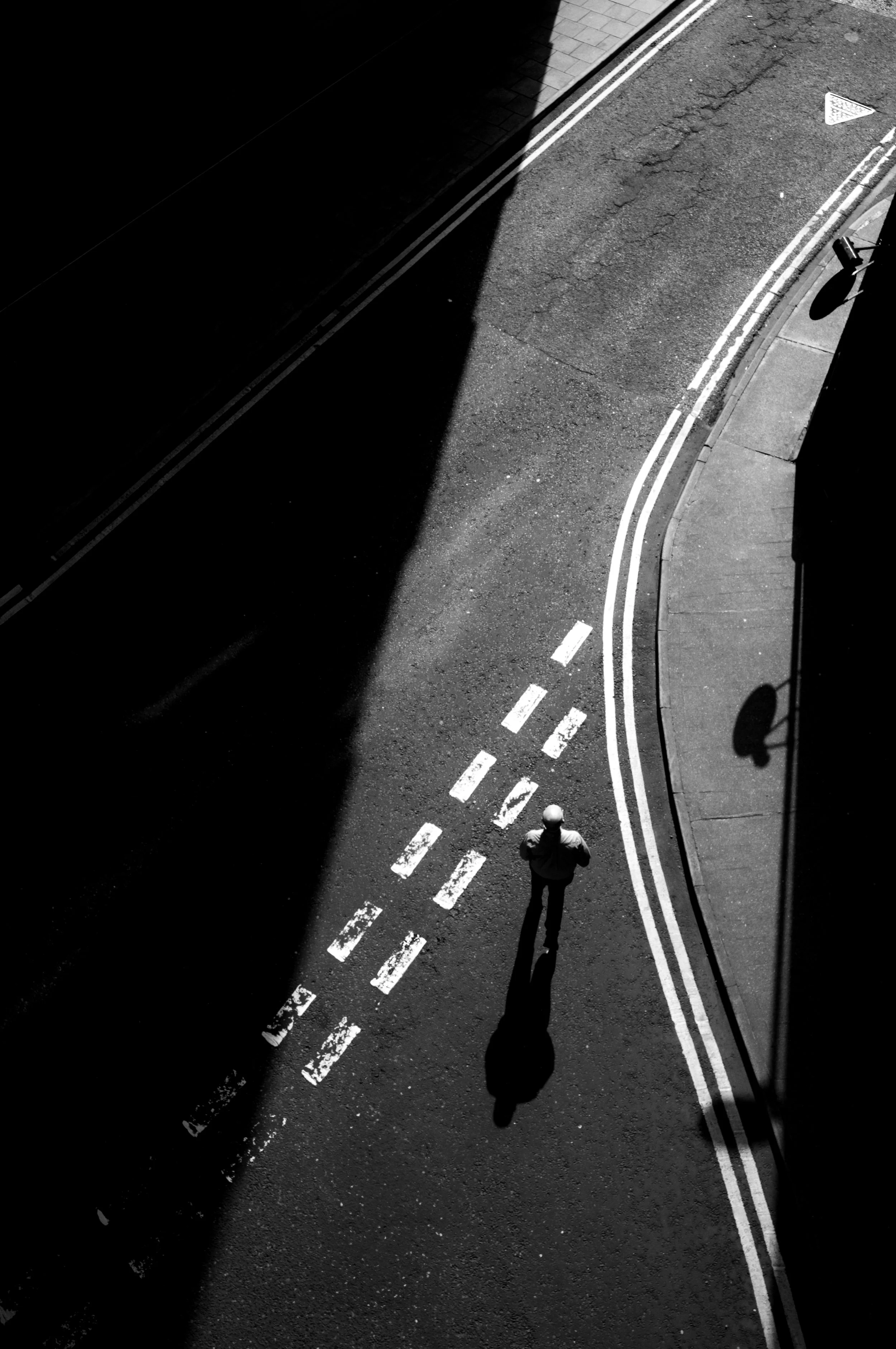 A black and white photo of a street corner, showing a person walking with a long shadow on the pavement, shadow cast by bright sunlight, with double traffic lines and street markings visible.