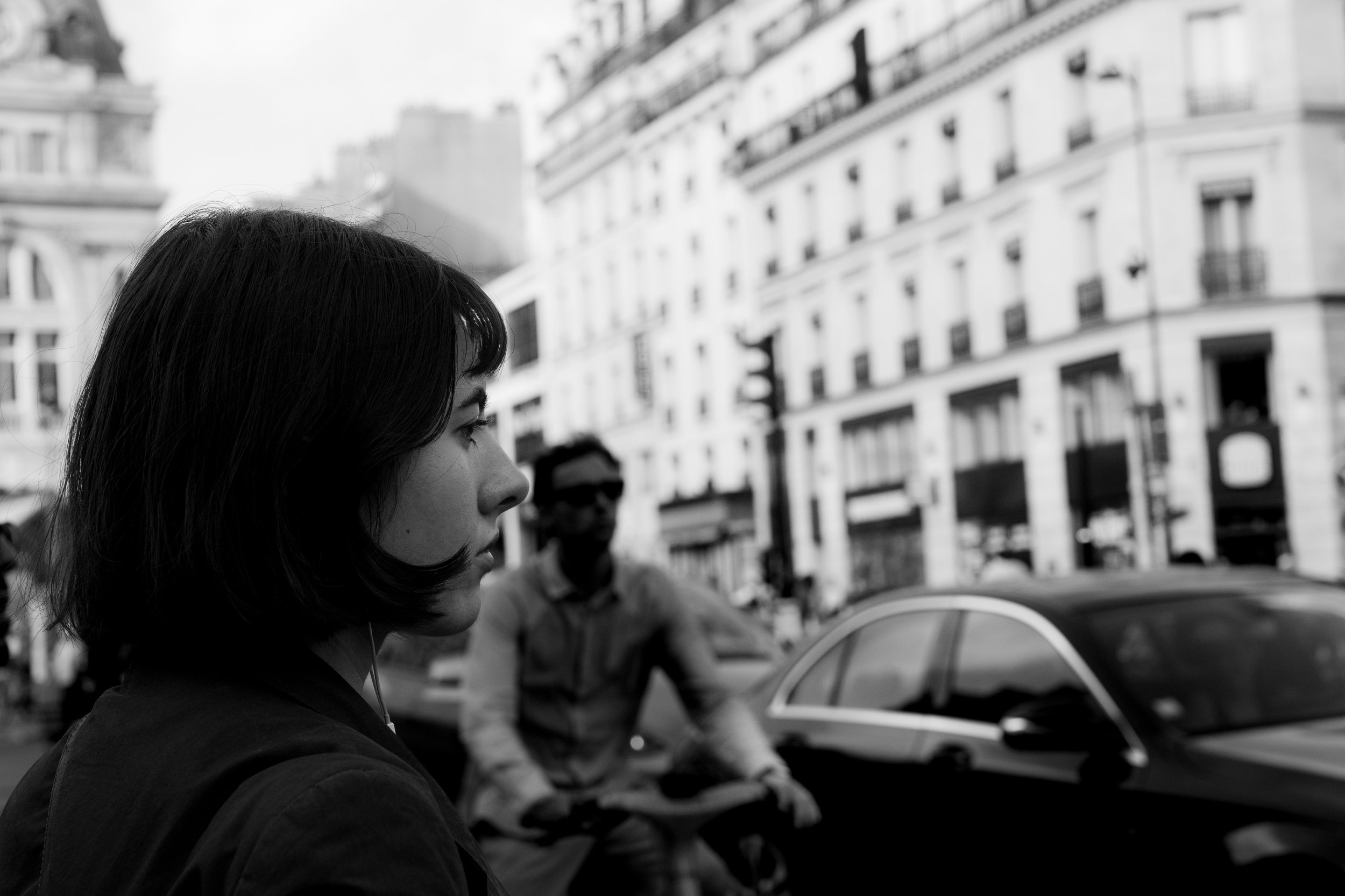Black and white photo of a woman with dark hair and earphones, standing on a busy city street with buildings, cars, and a person in the background.