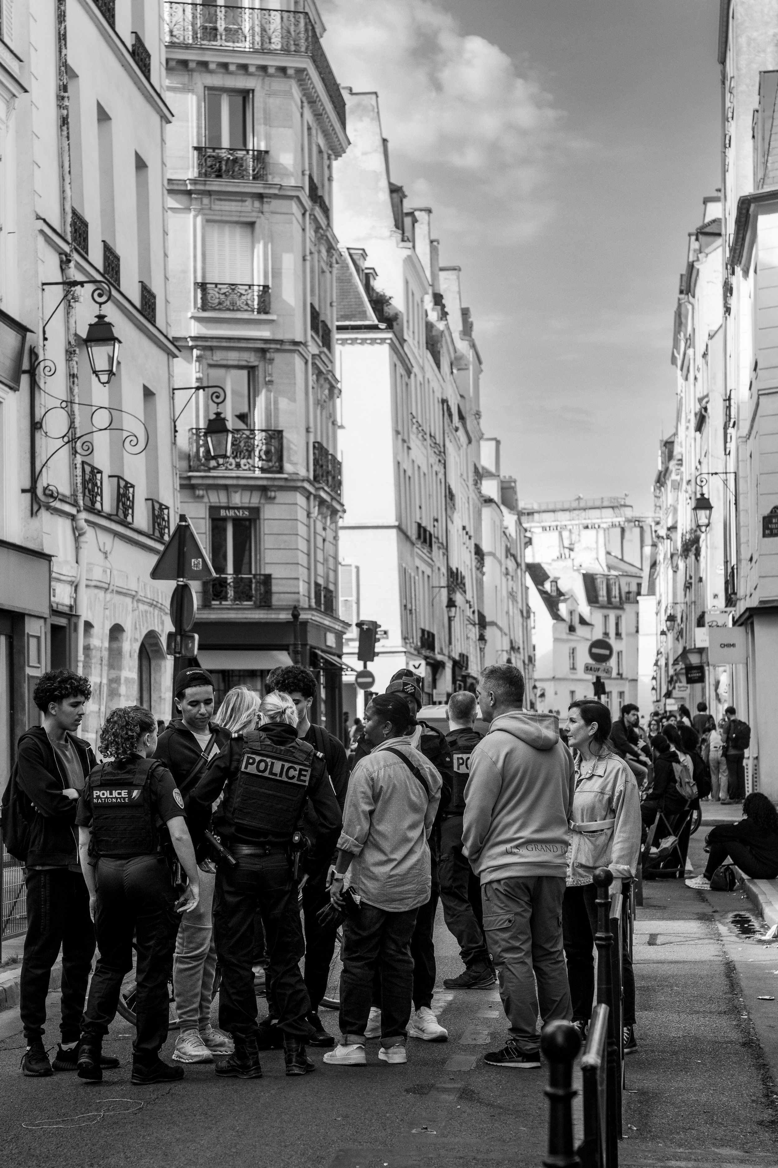 Group of police officers and bystanders talking on a street in a European city, with tall buildings, balconies, and street signs visible.