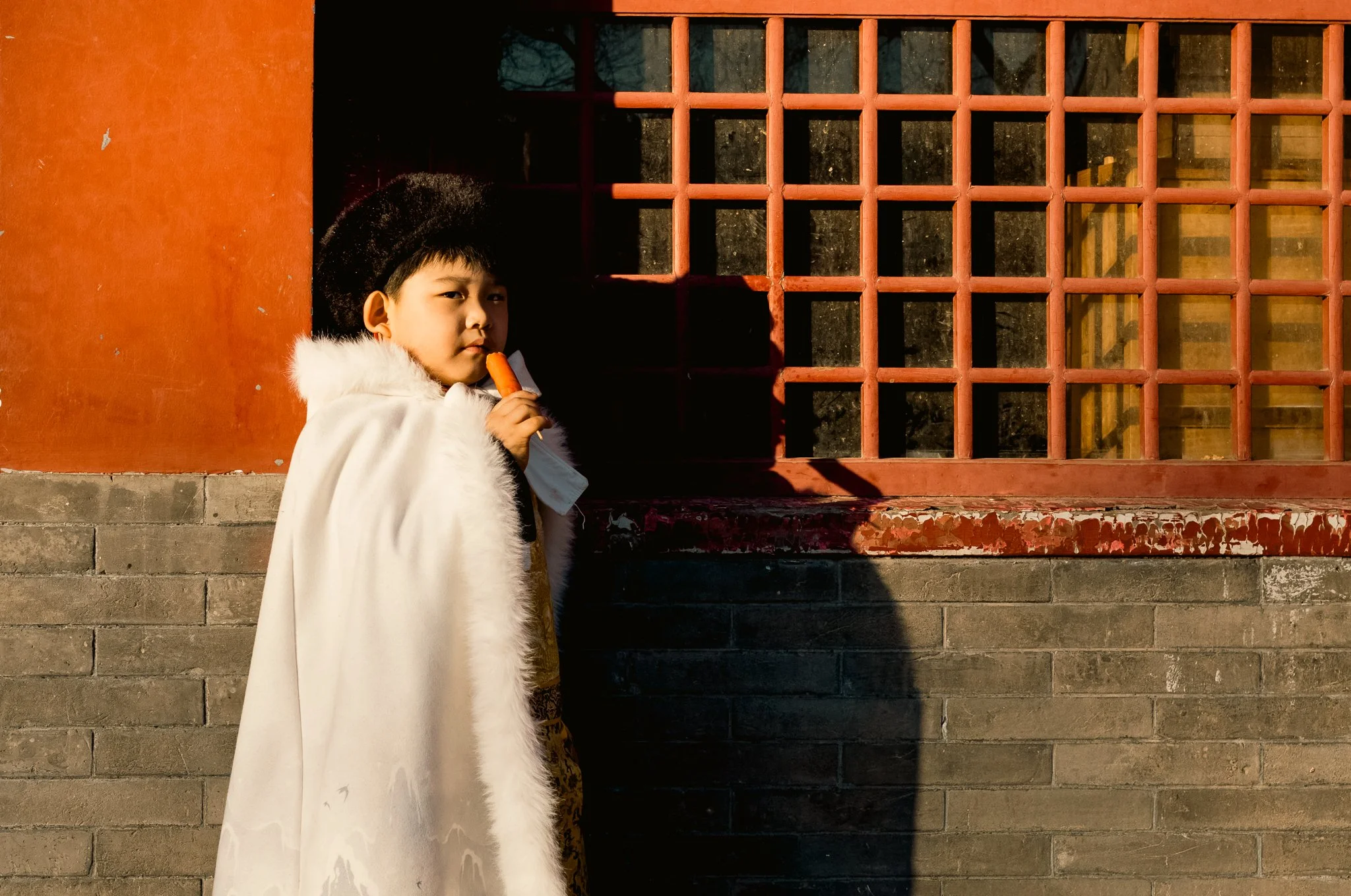 A young boy in traditional attire, wearing a white fur cloak and a large black hat, is standing against a brick wall next to a red window grate, holding a peeled carrot to his mouth.