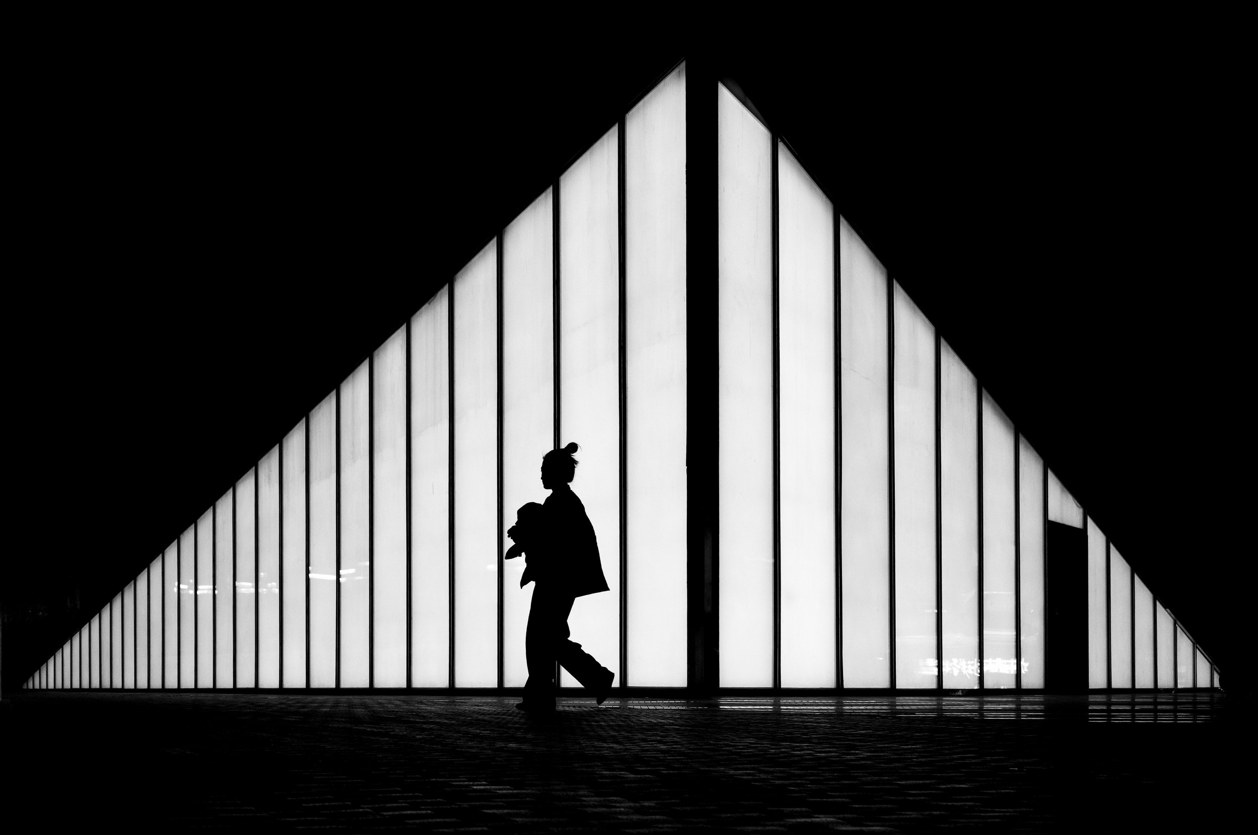 A silhouette of a person walking with a bag in front of a modern building with large glass windows.